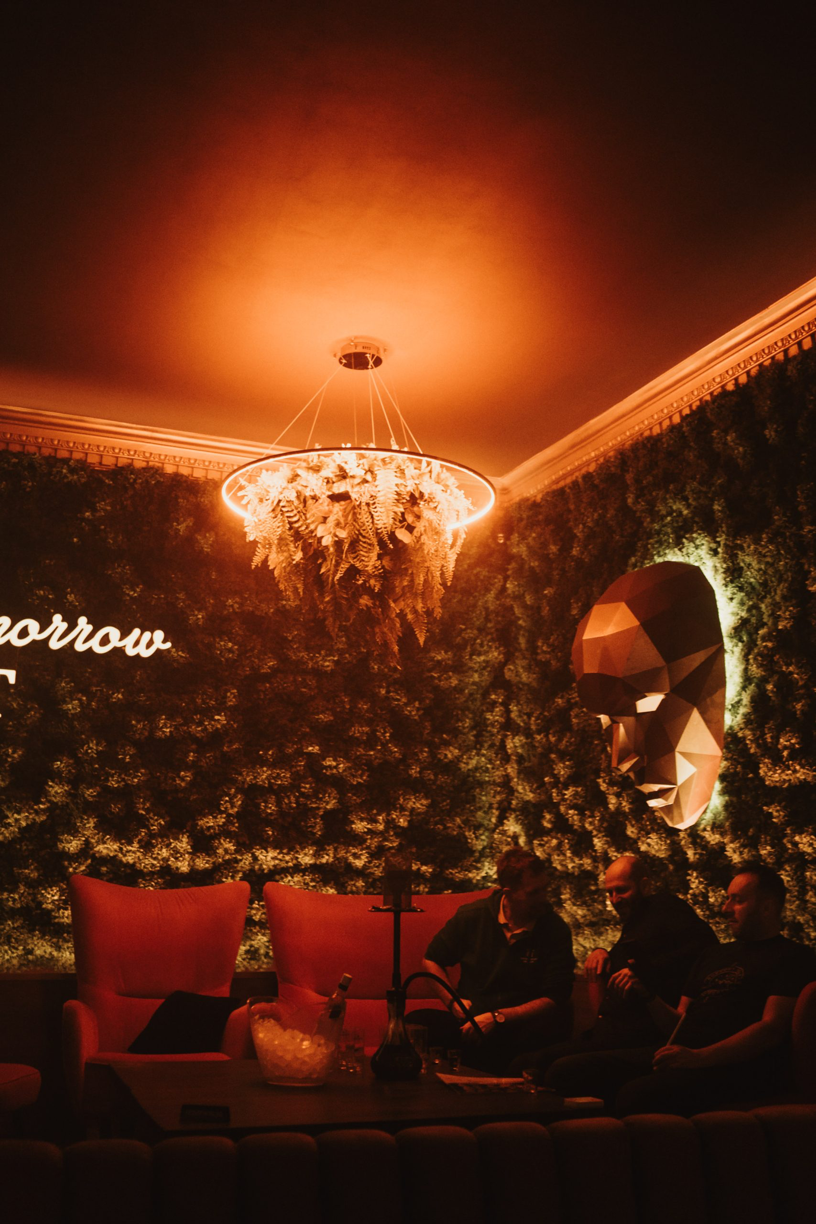 A stylish lounge area at Maska Incognito featuring a lush green wall, a large geometric face sculpture, and a circular chandelier with hanging ferns above a group of people enjoying a hookah.
