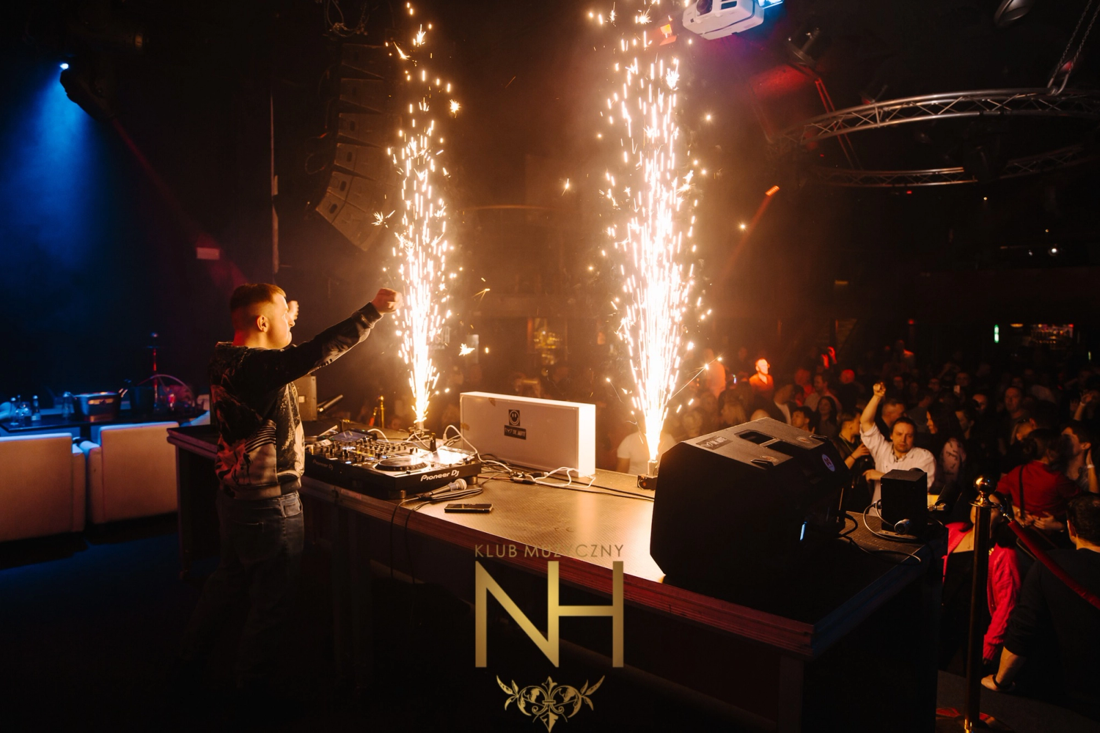 A DJ stands at a professional mixing console with two indoor sparkler fountains erupting in front of him, creating a bright and energetic atmosphere.