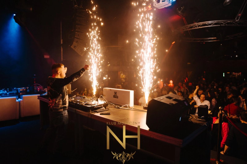 A DJ stands at a professional mixing console with two indoor sparkler fountains erupting in front of him, creating a bright and energetic atmosphere.
