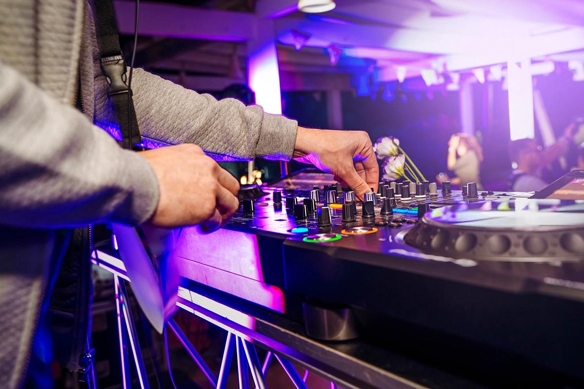 Close-up of a DJ's hands mixing tracks on a soundboard at a vibrant Warsaw rooftop club, illuminated by purple and blue party lights