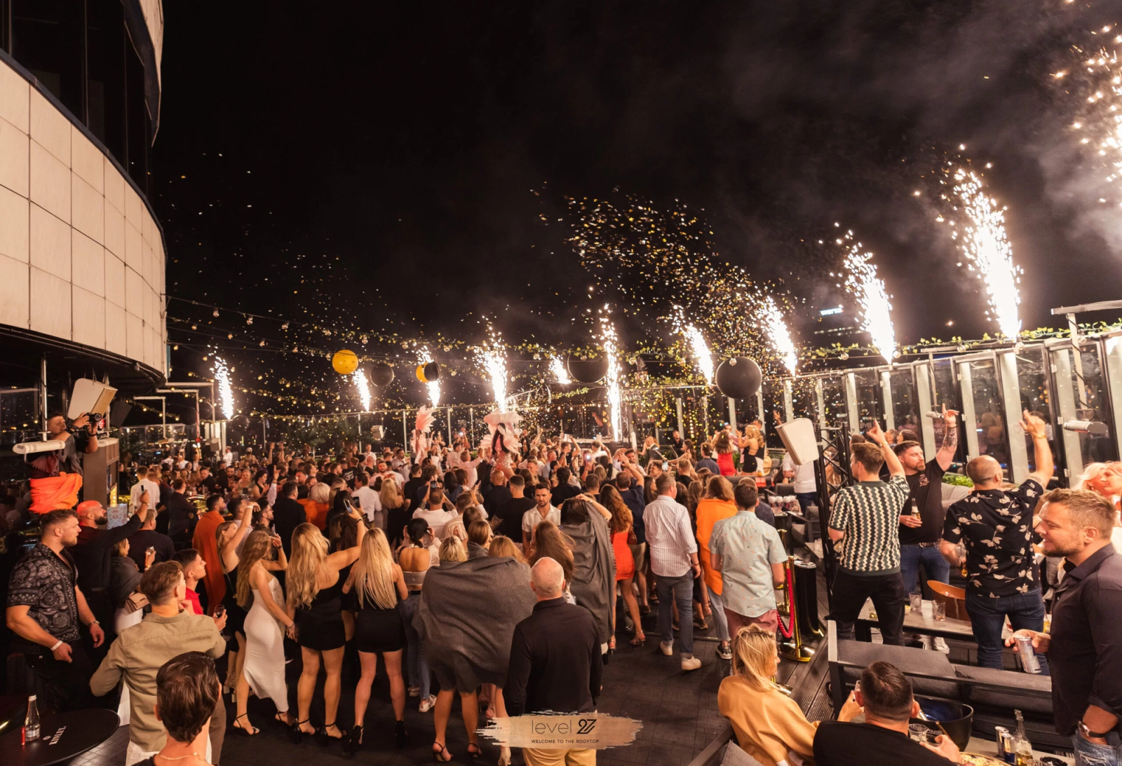 A vibrant night scene at the level 27 rooftop terrace in Warsaw, showing a large crowd of people dancing outdoors with sparkler fountains erupting along the glass perimeter against the city skyline.