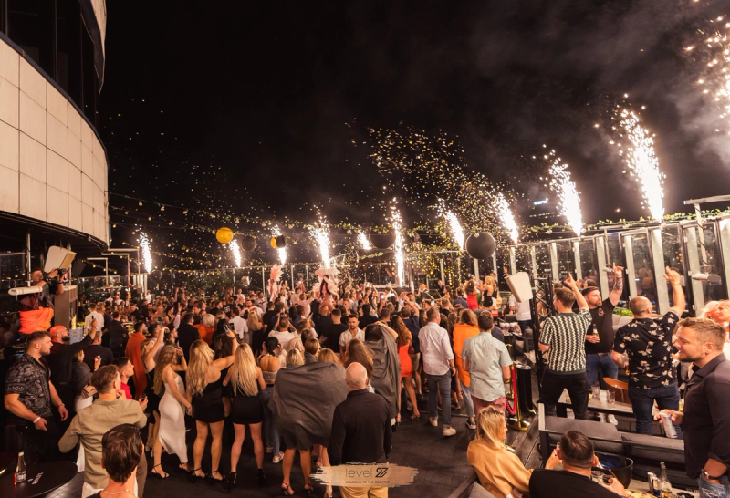 A vibrant night scene at the level 27 rooftop terrace in Warsaw, showing a large crowd of people dancing outdoors with sparkler fountains erupting along the glass perimeter against the city skyline.