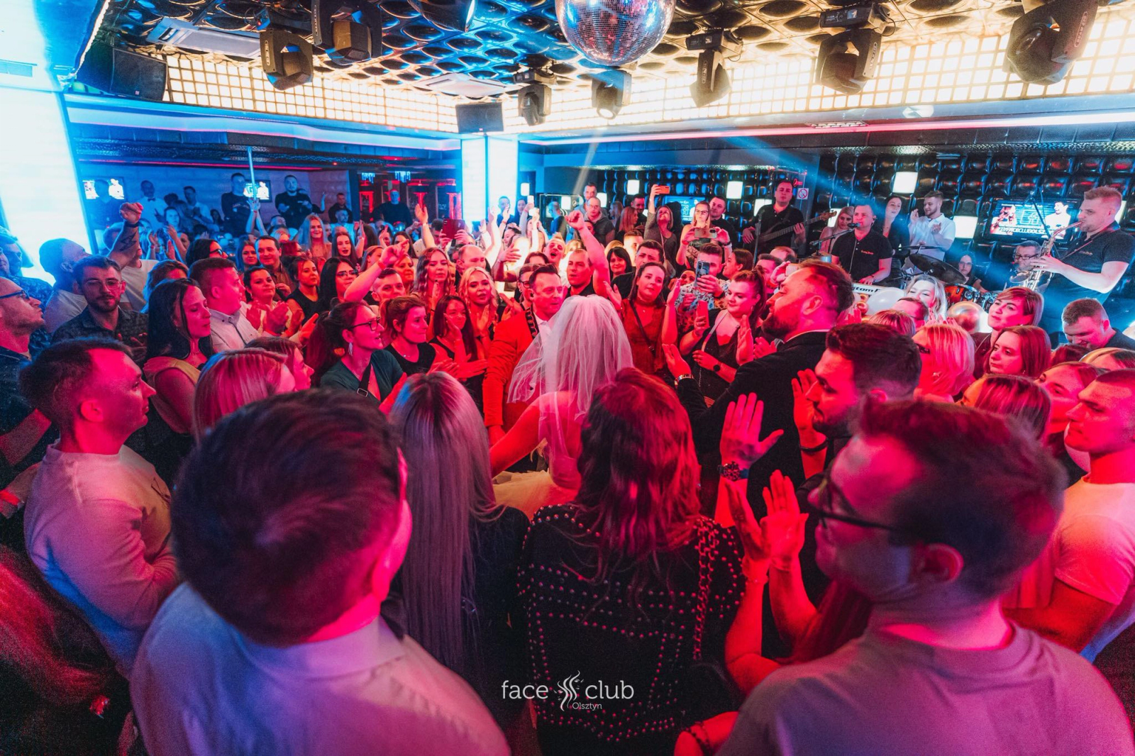 A dense crowd on the dance floor at Face Club Olsztyn, featuring a bride in a veil and a groom in a red jacket celebrating in the center under disco balls and spotlights.