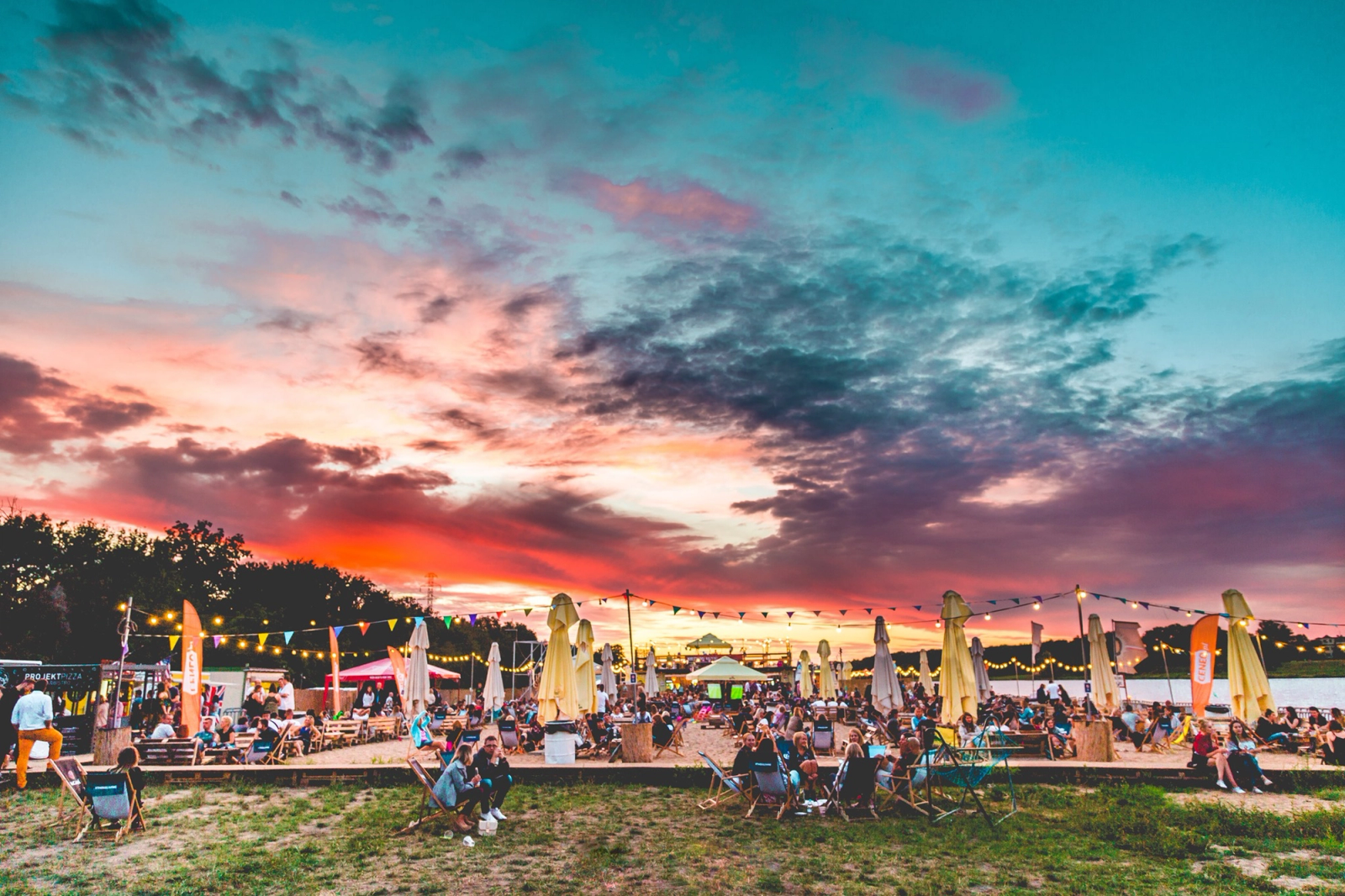 A breathtaking panoramic view of HotSpot BeachBar at sunset, with a vibrant orange and purple sky reflecting over a sandy area filled with people, deck chairs, and festive string lights.