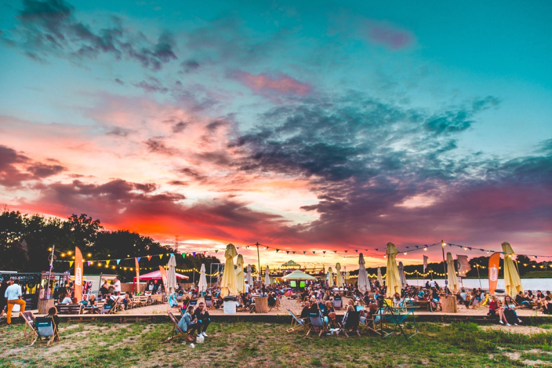 A breathtaking panoramic view of HotSpot BeachBar at sunset, with a vibrant orange and purple sky reflecting over a sandy area filled with people, deck chairs, and festive string lights.