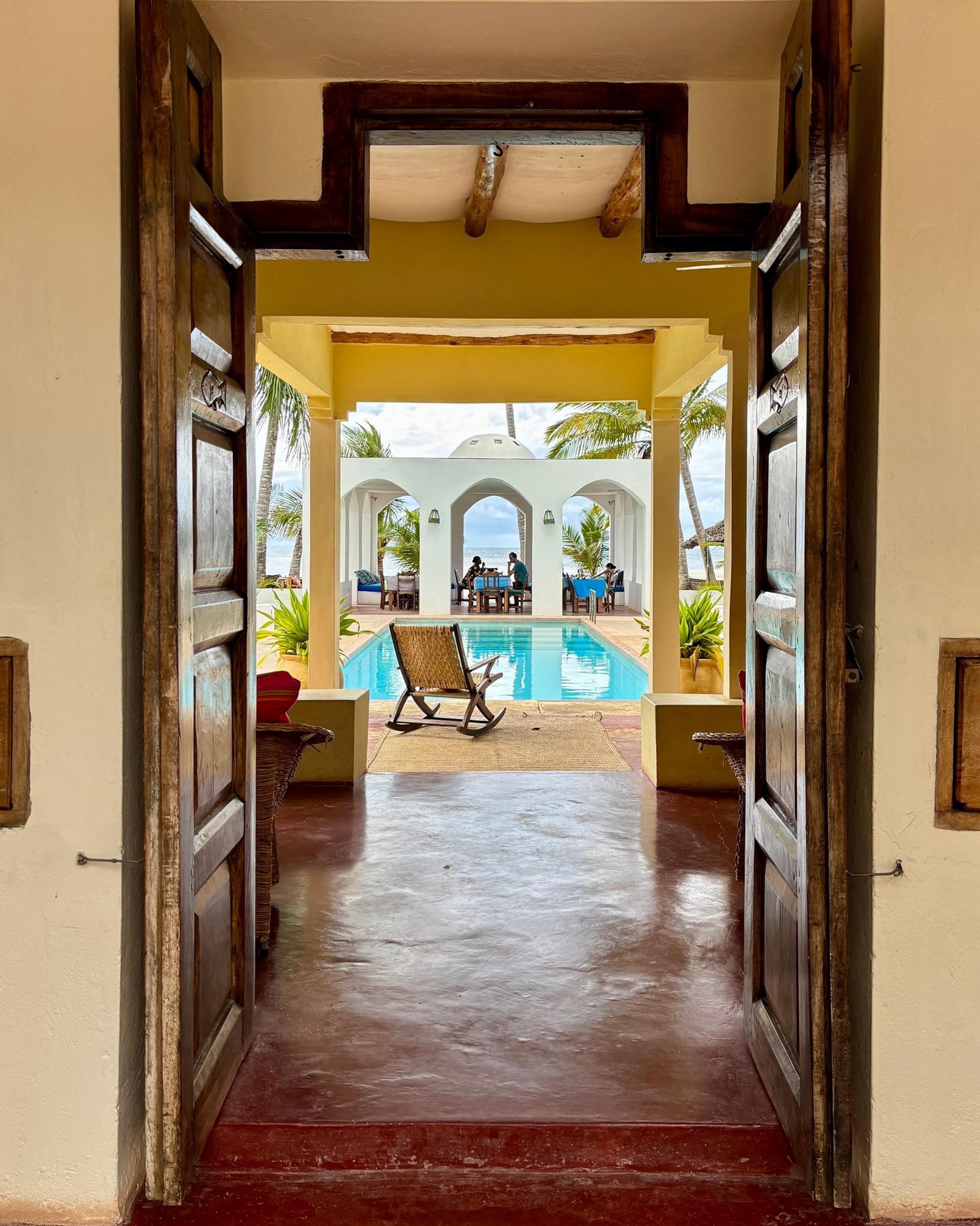 A carved timber doorway framing the arched pool and ocean view at Sawa Sawa Beach House