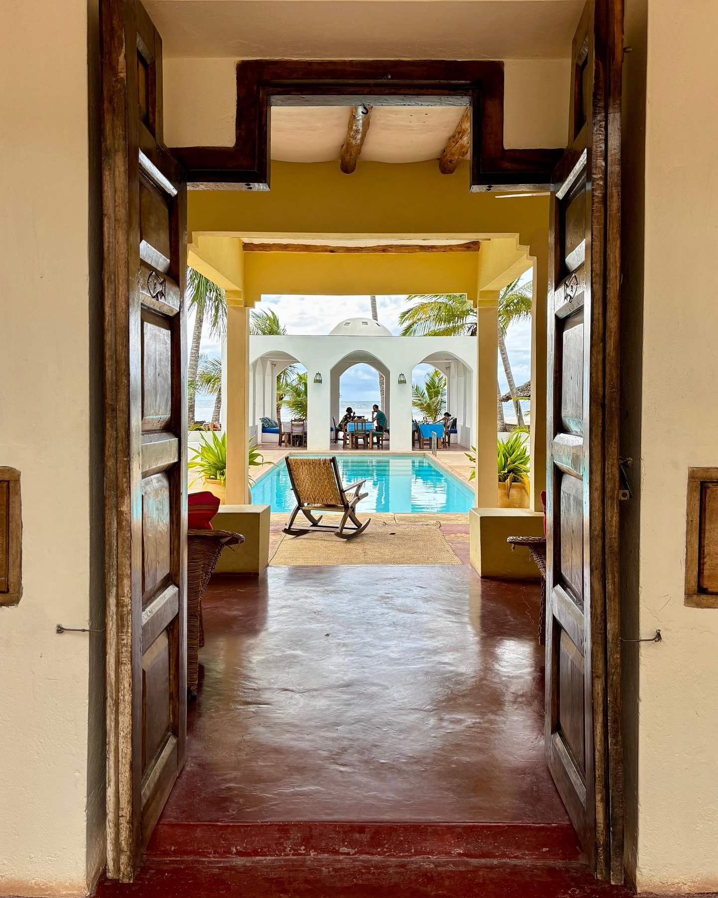 A carved timber doorway framing the arched pool and ocean view at Sawa Sawa Beach House