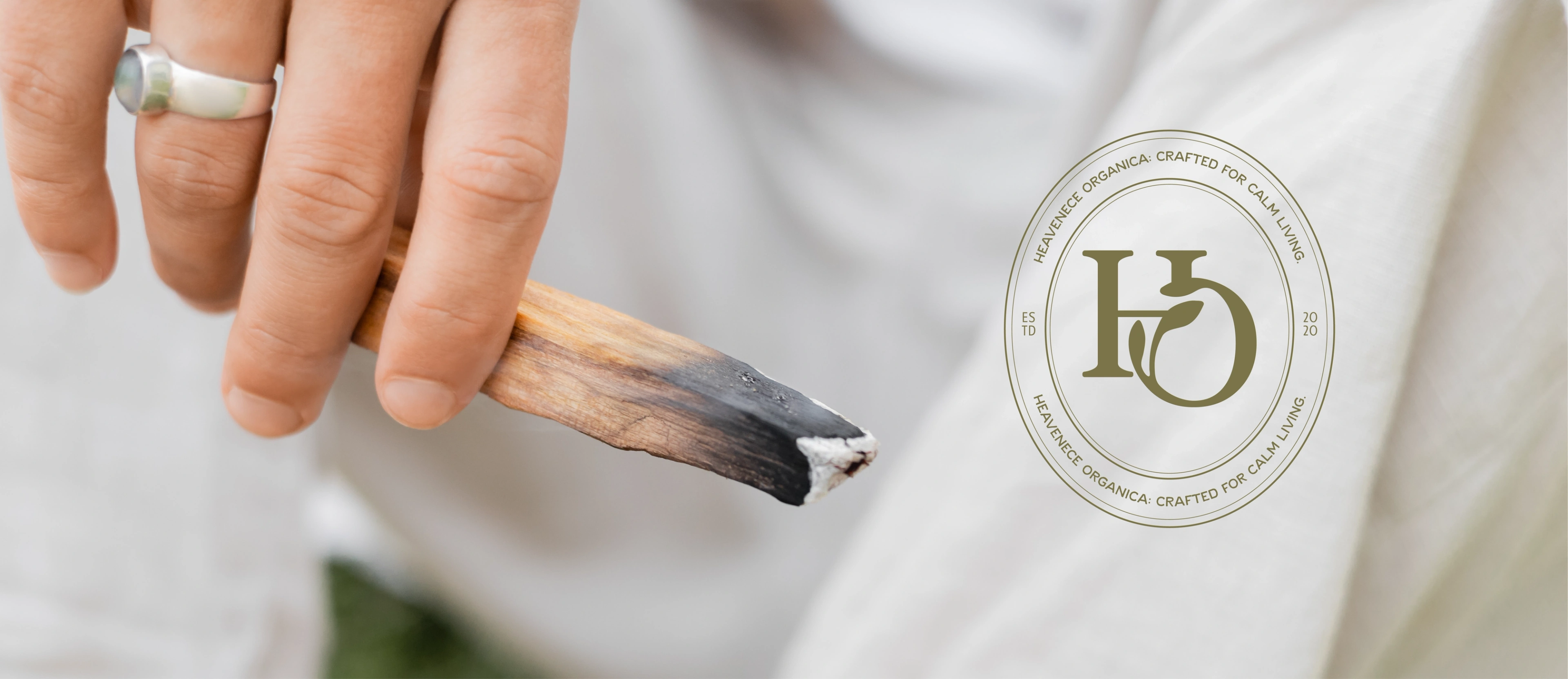 A hand holding a smouldering Heavenence Organica incense stick beside the HO seal