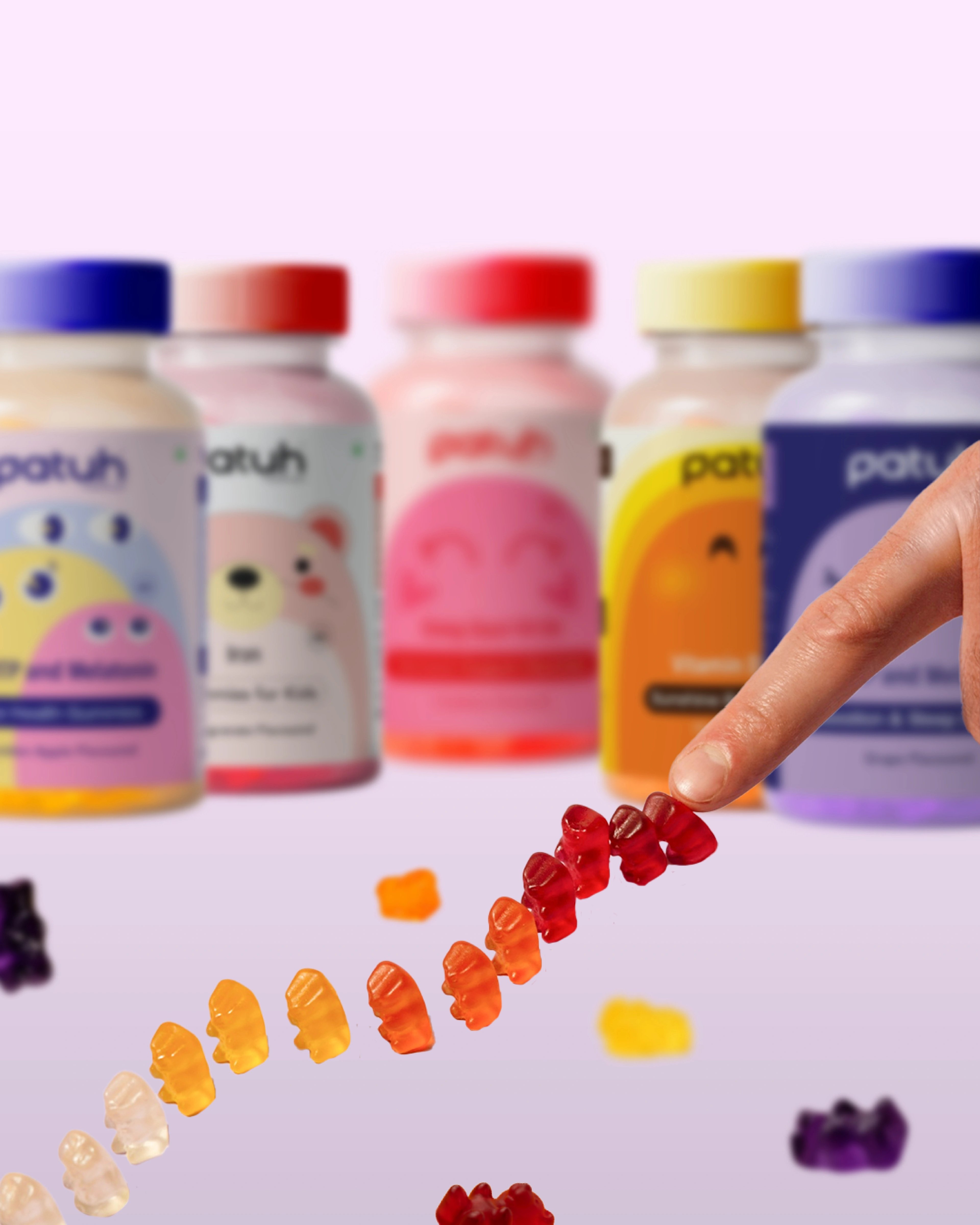 A hand reaching toward an arc of Patuh gummy bears in flavour-graded colours, the full product range blurred behind