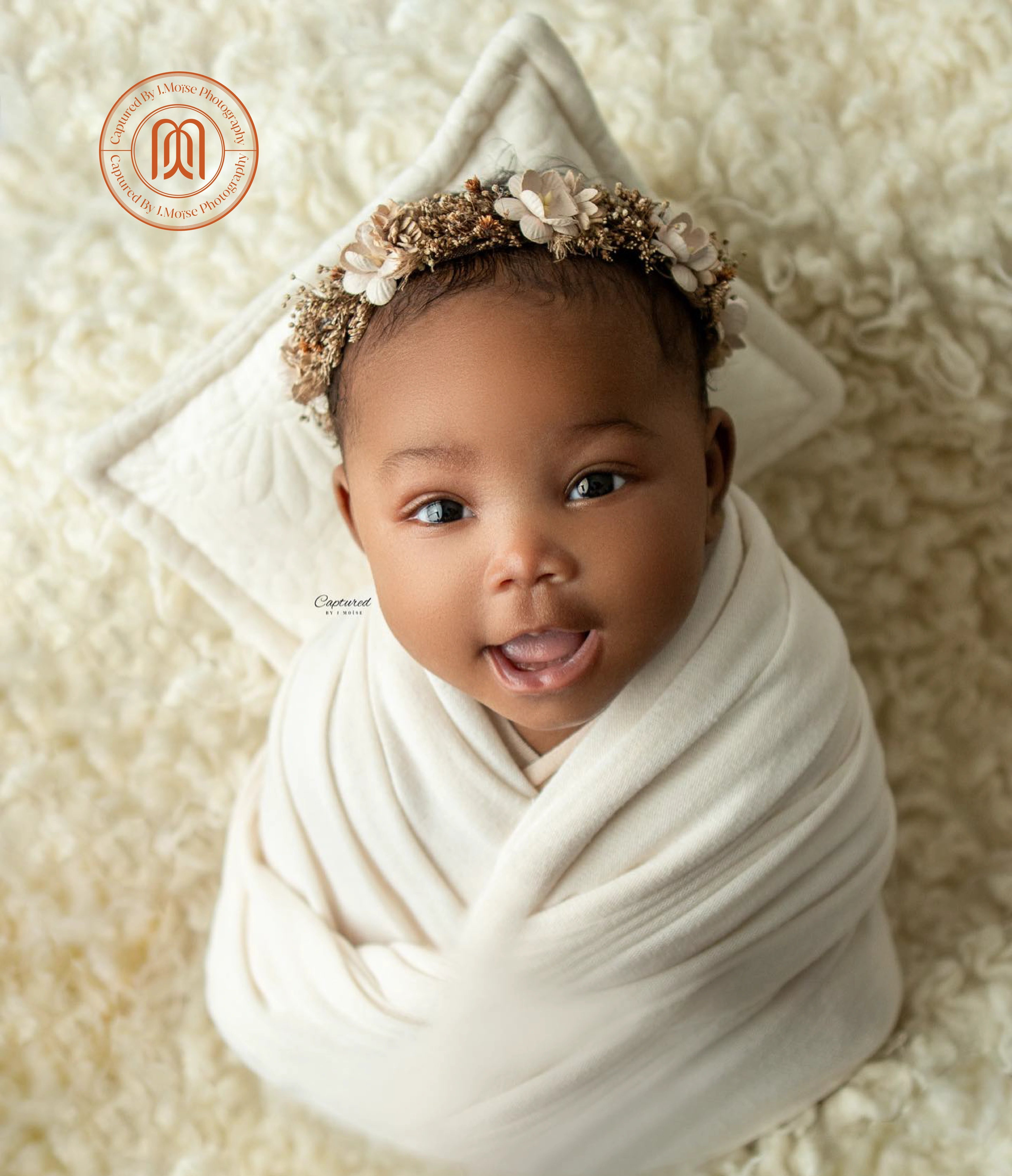 a newborn swaddled in cream lying on soft fur, the studio monogram in the corner
