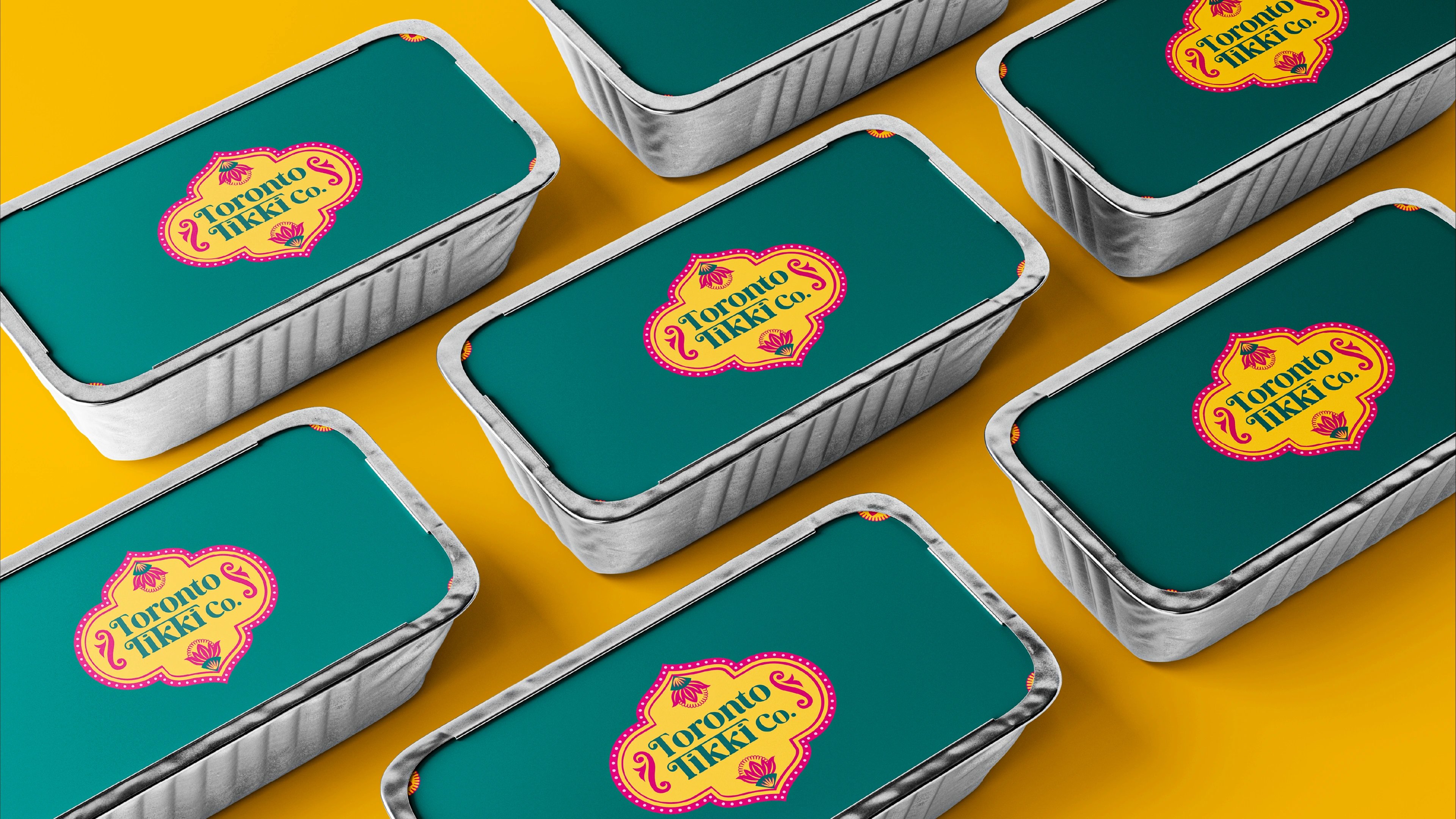 Teal Toronto Tikki Co. takeout tins with lotus-crest labels shot top-down on a yellow surface