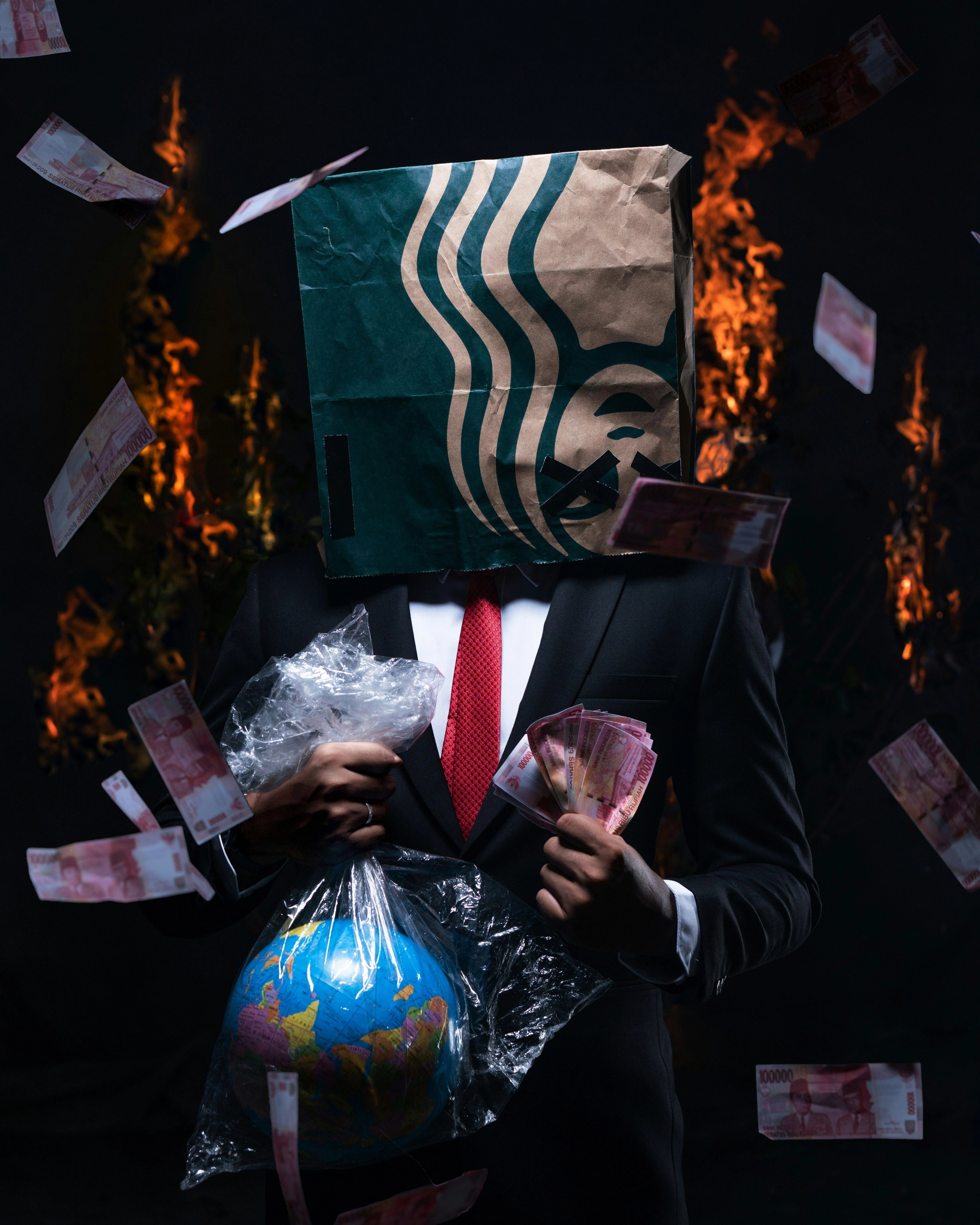 A conceptual photo of a suited man with a Starbucks paper bag over his head holding fistfuls of cash and a plastic-wrapped globe as banknotes burn around him