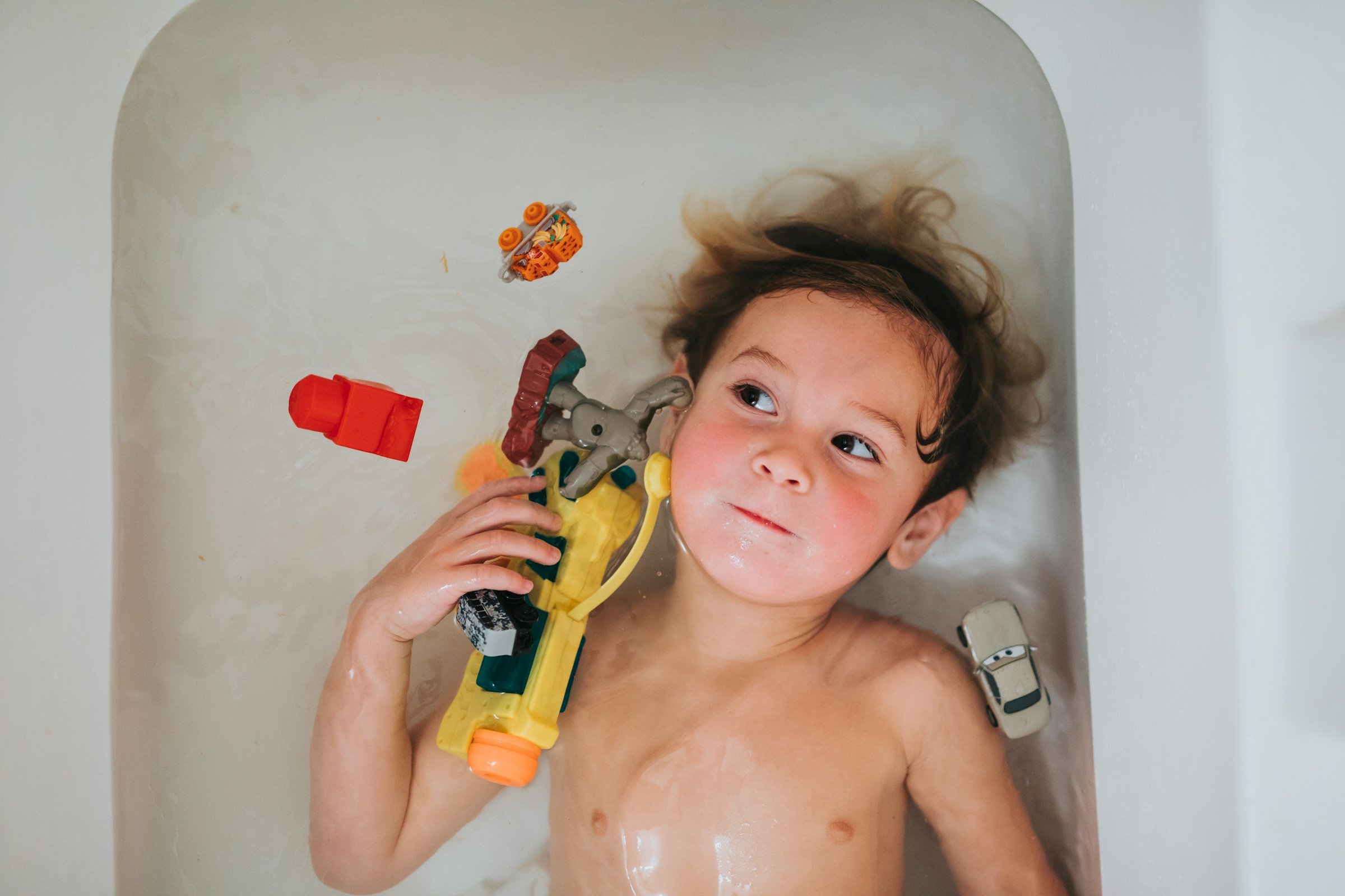A young child lying in a bathtub at play, clutching a yellow toy water-blaster with a toy car resting on their shoulder