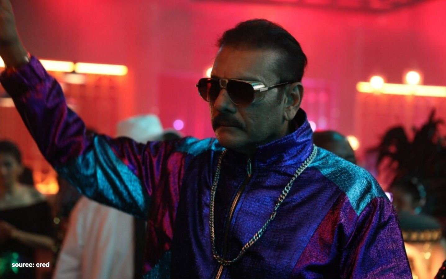 A still from a CRED ad — a retro-styled man in a shiny metallic jacket, aviators and gold chains raises his arm on a neon party floor
