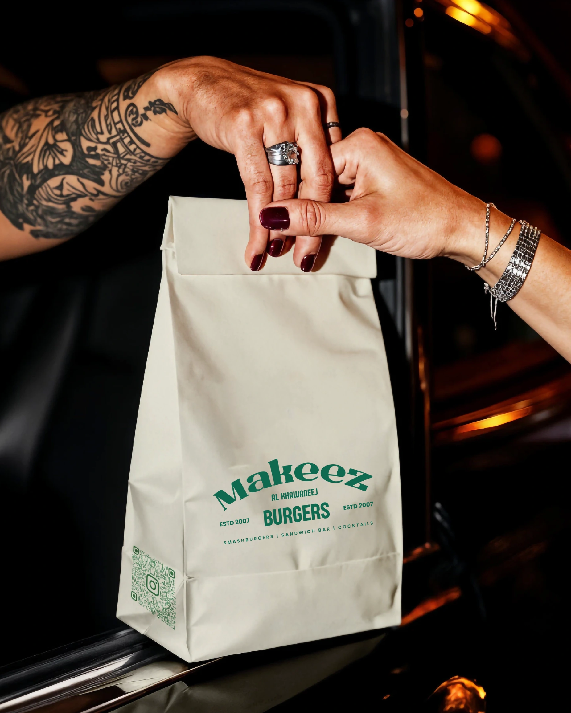 Gloved hands holding a Makeez burger beside branded takeaway bags