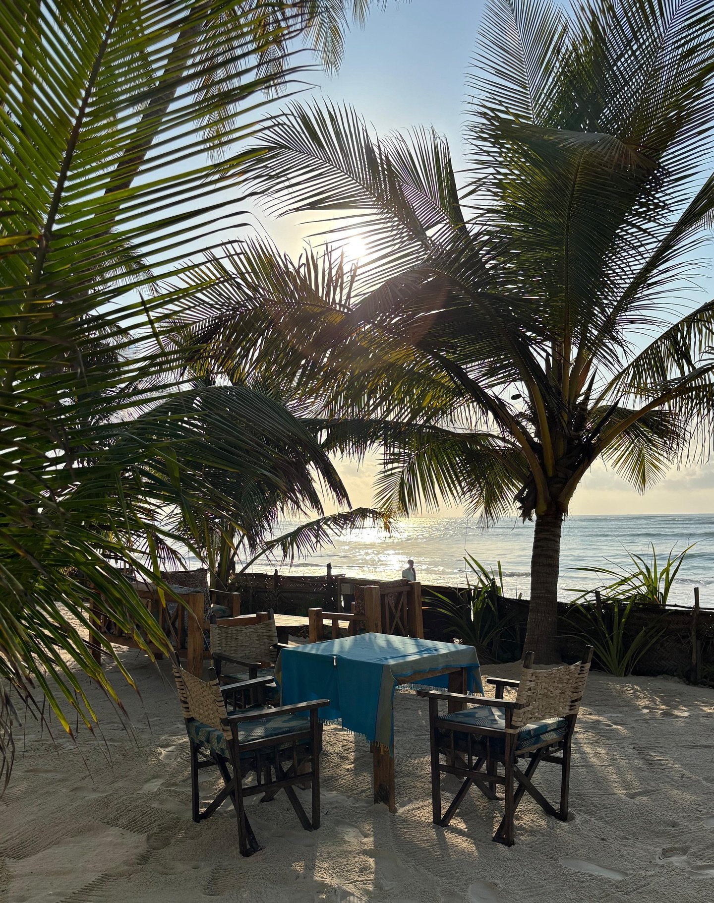 A driftwood table and chairs on the sand under palms at Sawa Sawa Beach House, ocean beyond at golden hour
