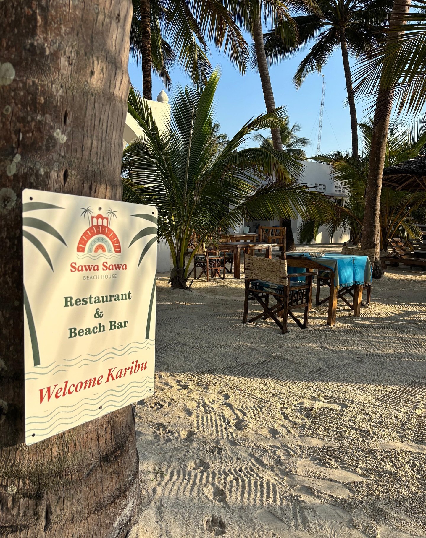 The Sawa Sawa 'Restaurant & Beach Bar — Welcome Karibu' sign fixed to a palm trunk on the beach