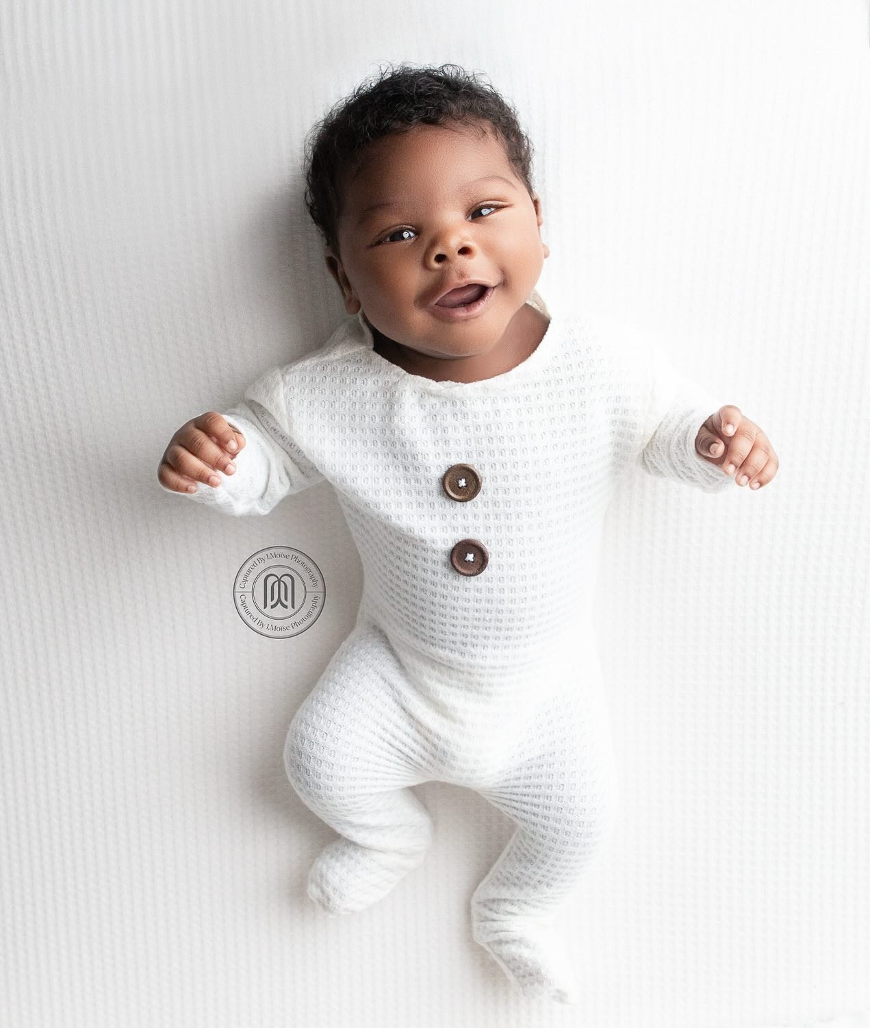 a Captured by J. Moïse newborn portrait — a smiling baby in a white waffle-knit onesie on a white blanket, the studio seal watermark centred