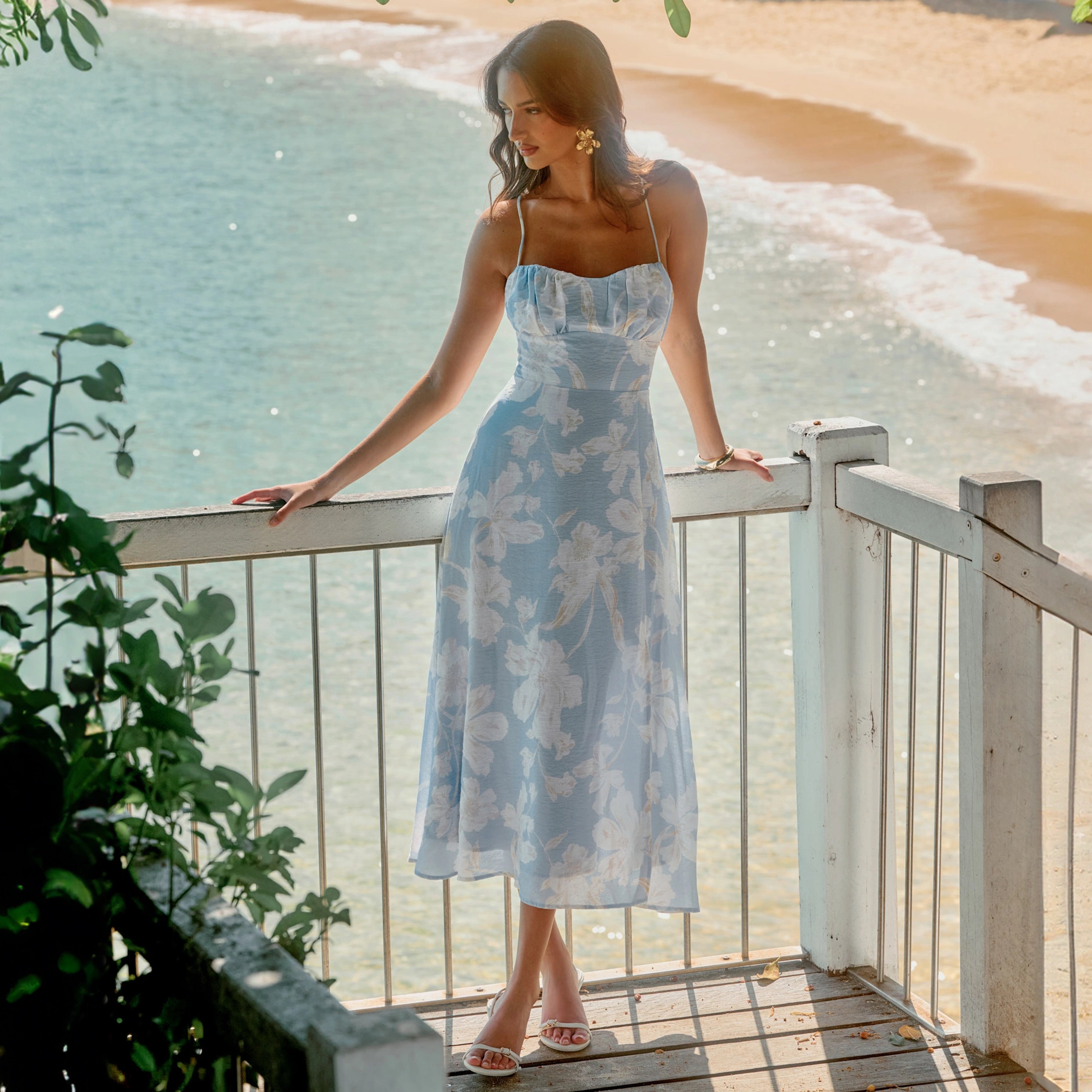 A coastal fashion frame by Elyse Potter — a model in a pale-blue floral dress leaning on a white balcony rail above a sunlit cove