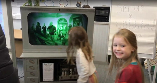 Two children one smiling at the camera, the other looking away, in front of a green tinted reflection shown on a screen in a grey cabinet.