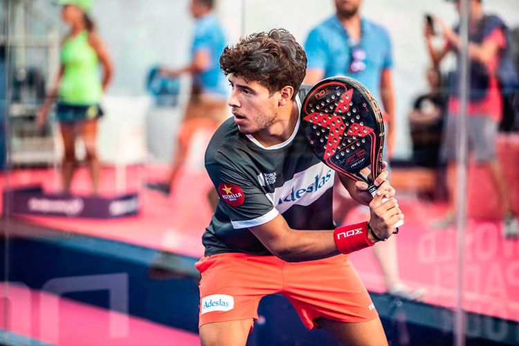 Agustín Tapia waits for the ball in the ready position, racket raised, on a glass-walled padel court