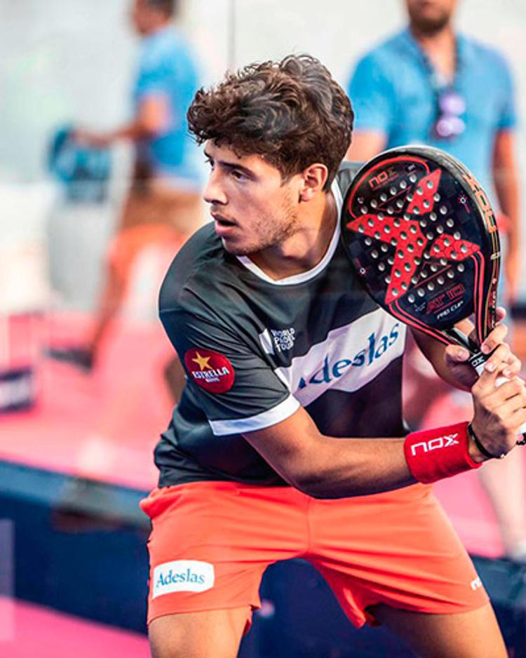 Agustín Tapia waits for the ball in the ready position, racket raised, on a glass-walled padel court