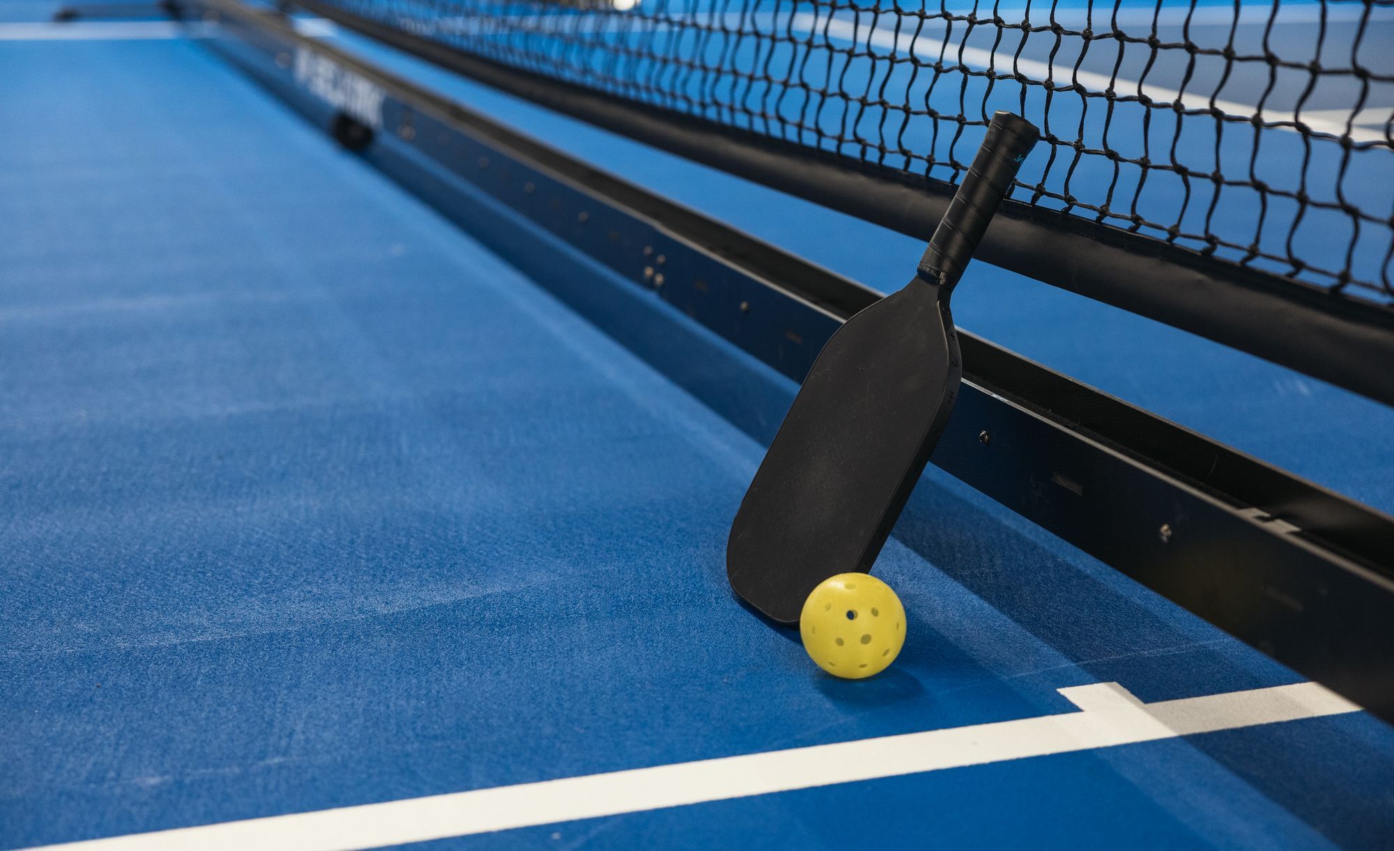 A portable pickleball net on a blue court, with a paddle and yellow holed ball resting against its frame