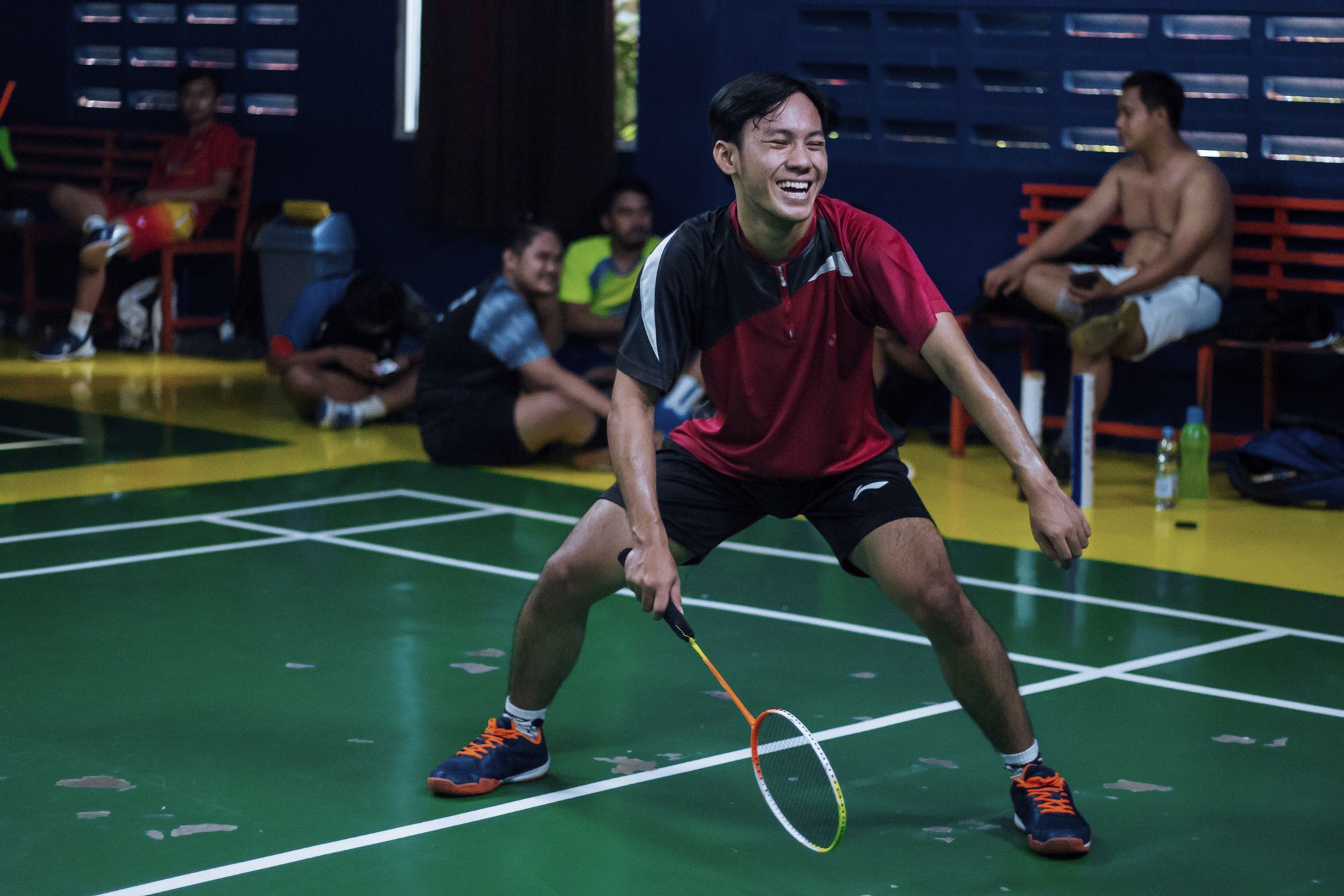 A badminton player in a ready stance on a green indoor court, with other players resting at the side of the hall