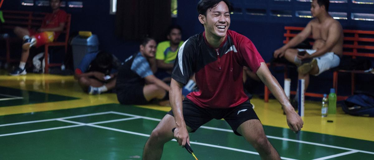 A badminton player in a ready stance on a green indoor court, with other players resting at the side of the hall