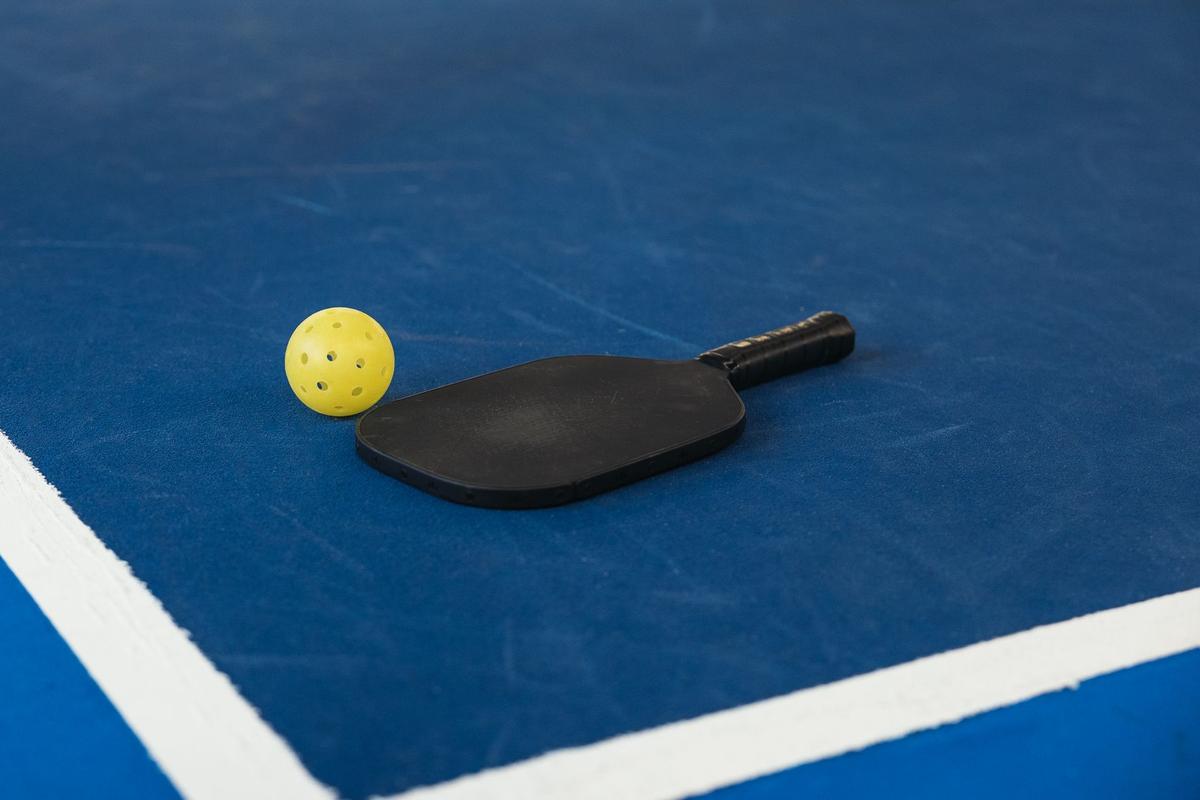A yellow perforated plastic pickleball ball beside a black paddle on a blue court near the white lines