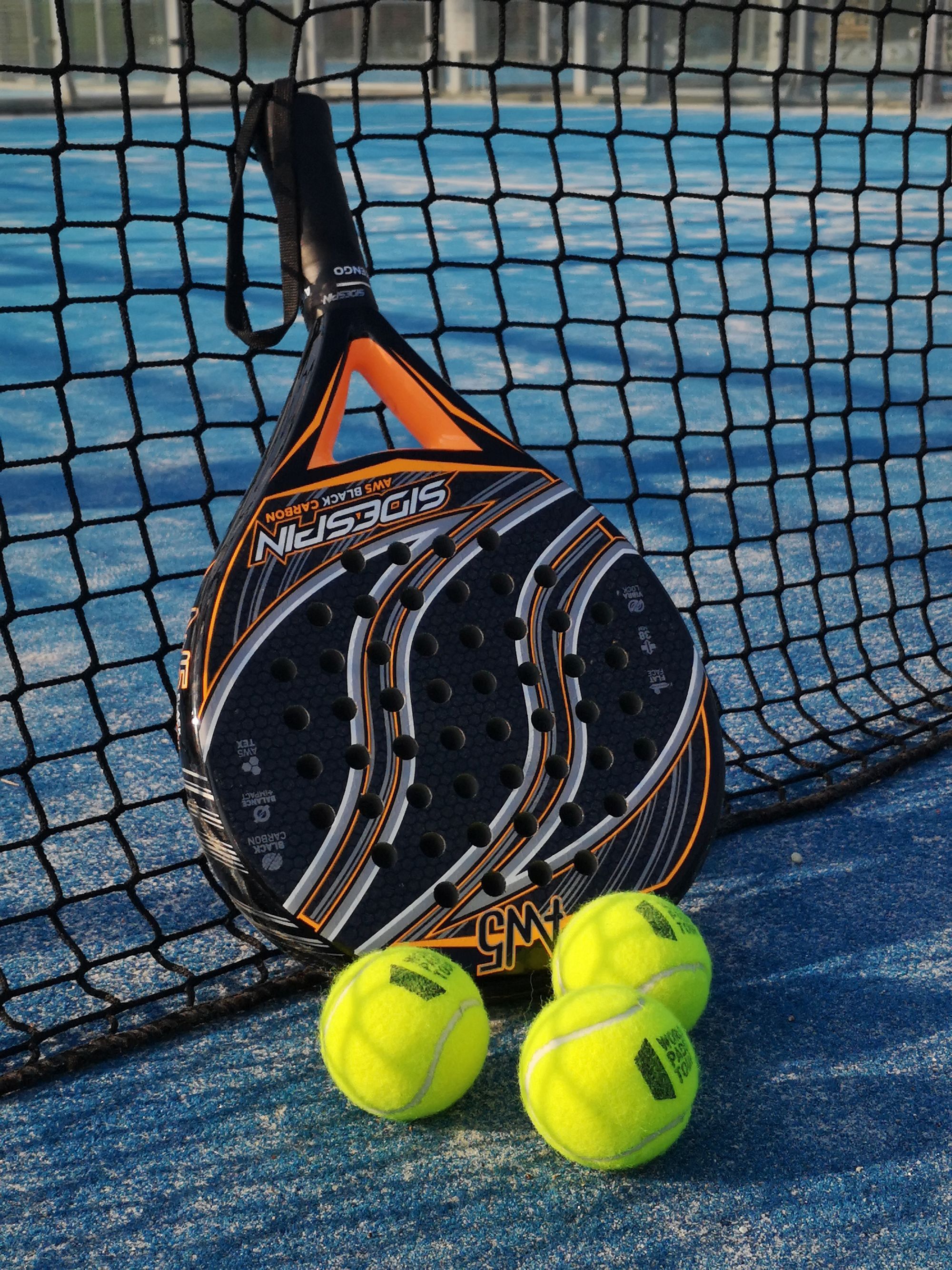 A solid, perforated padel racket leaning against the net on a blue outdoor court, with three padel balls at its base