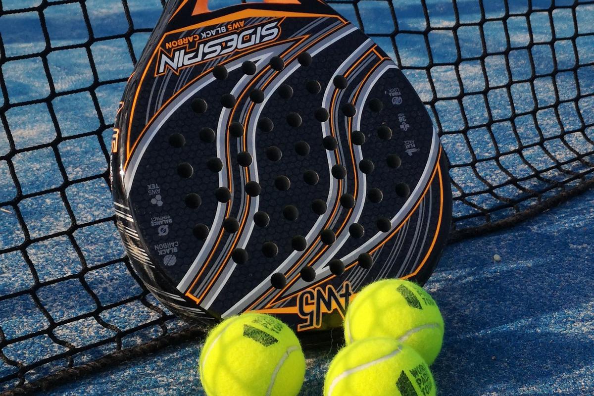 A solid, perforated padel racket leaning against the net on a blue outdoor court, with three padel balls at its base