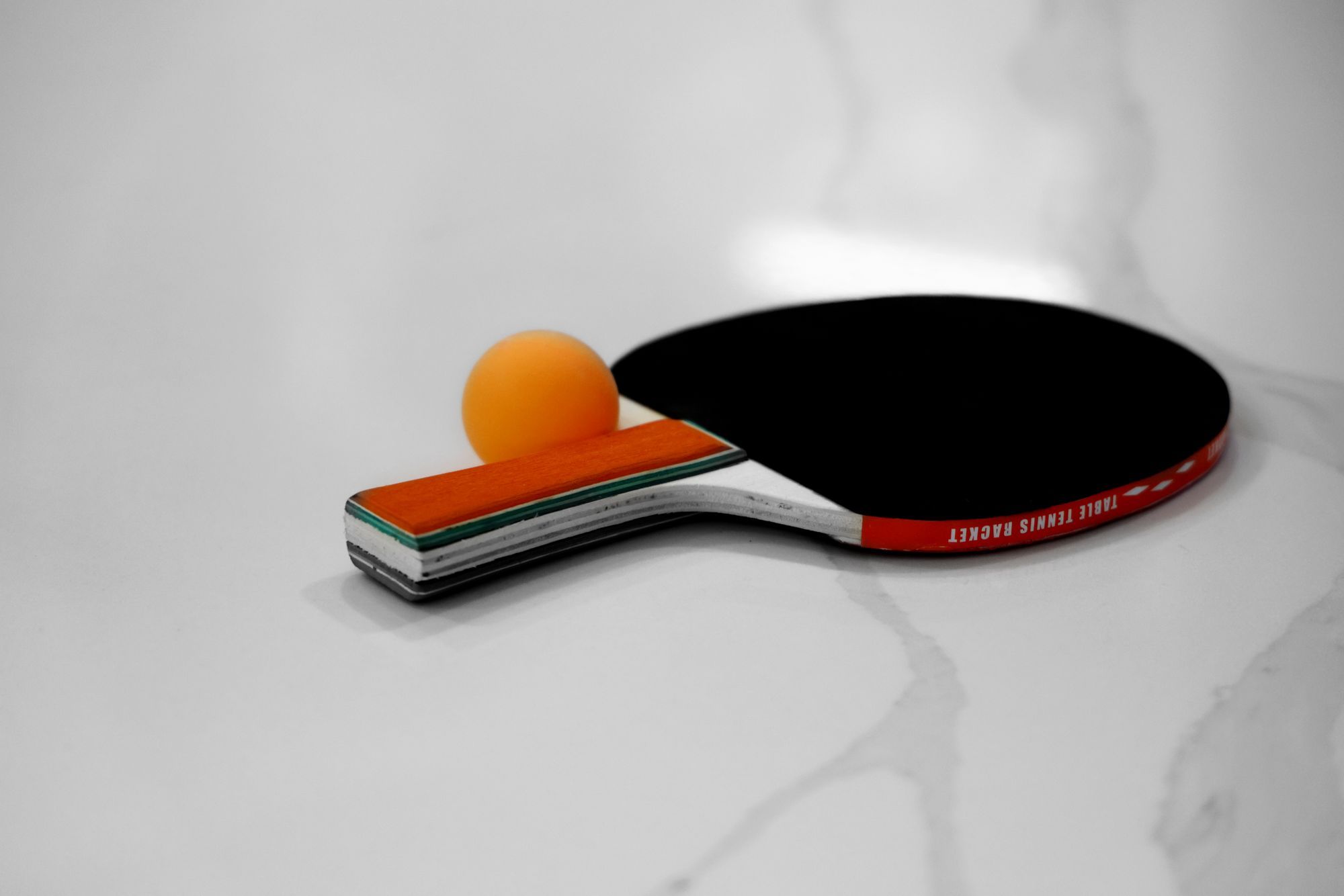 A table tennis bat with black rubber and a layered wooden handle lying on white marble beside an orange ball