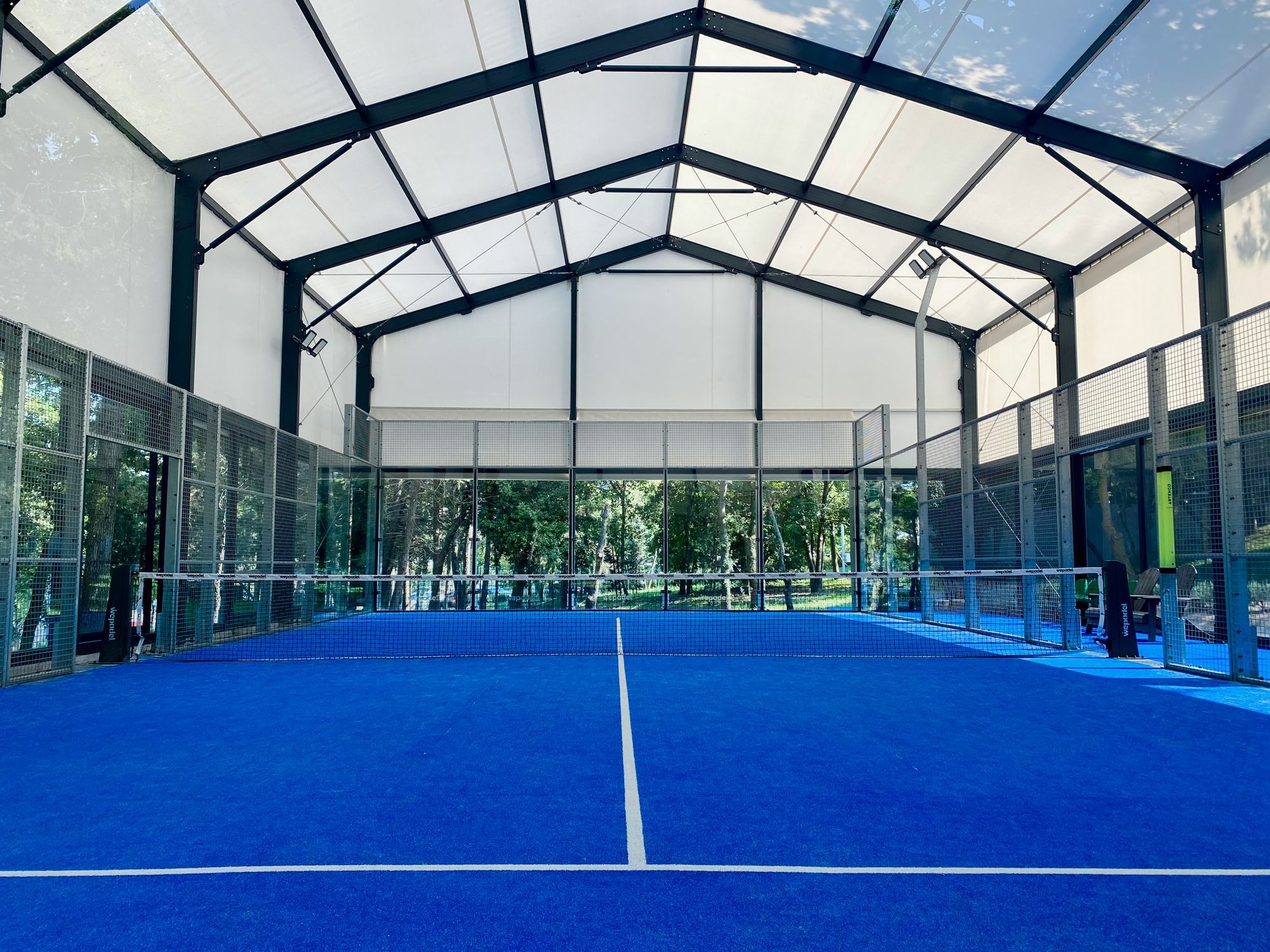The net across an indoor padel court with a blue surface, enclosed by glass back walls and mesh side fencing