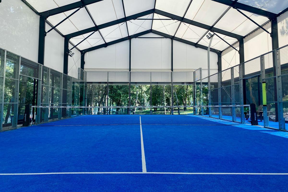 The net across an indoor padel court with a blue surface, enclosed by glass back walls and mesh side fencing
