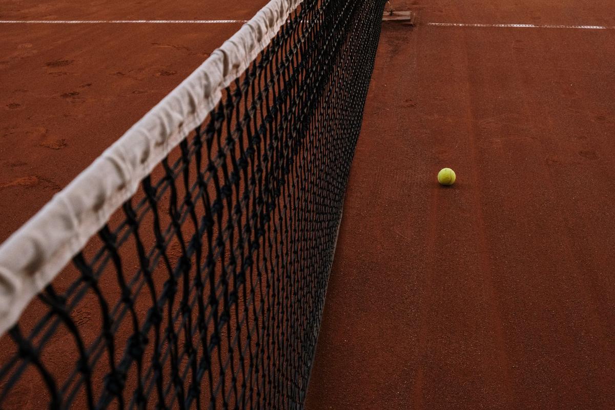 A tennis net with its white headband stretched across a clay court, with a single ball lying beyond it