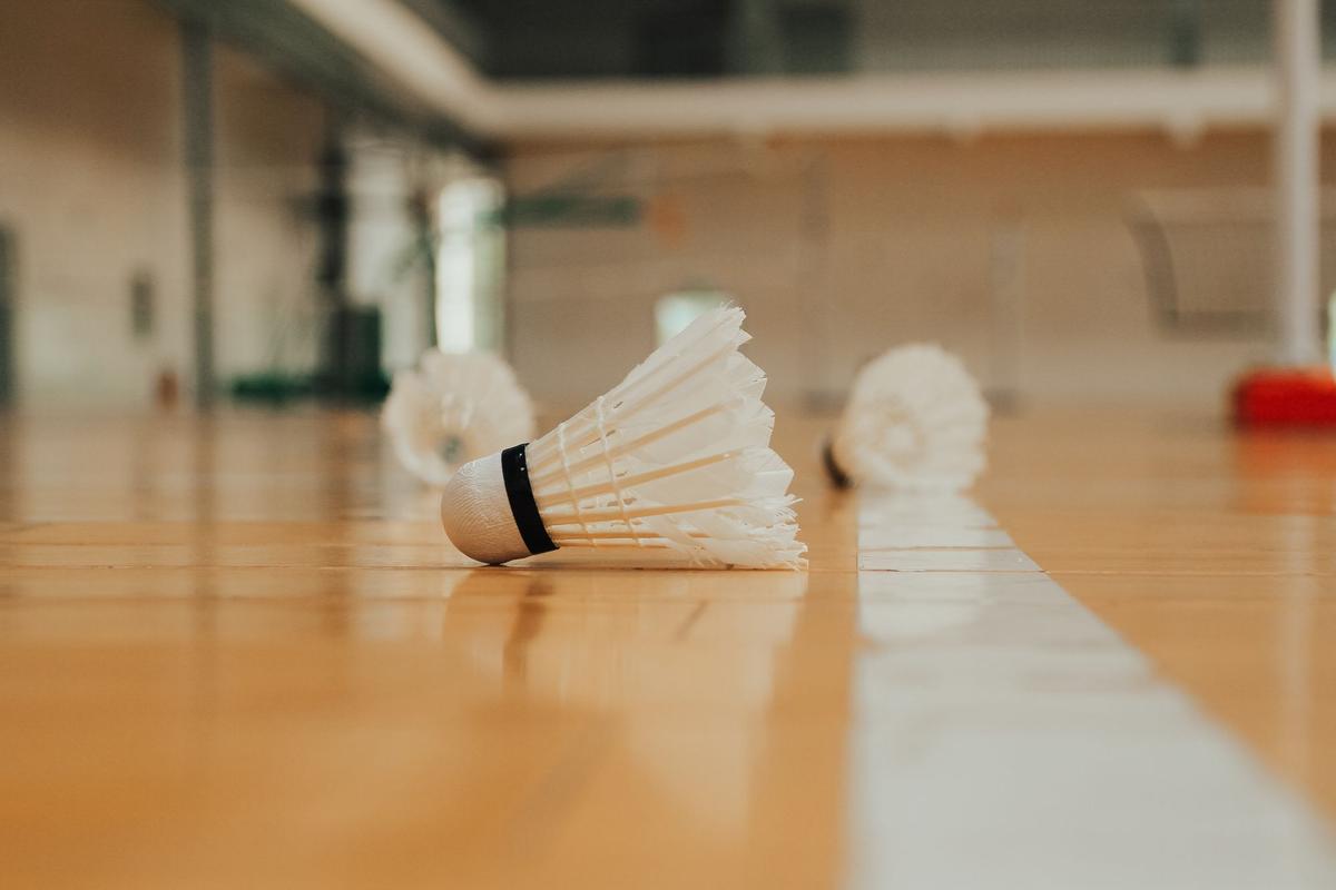 Feather badminton shuttlecocks with cork bases lying on a wooden sports hall floor beside a court line