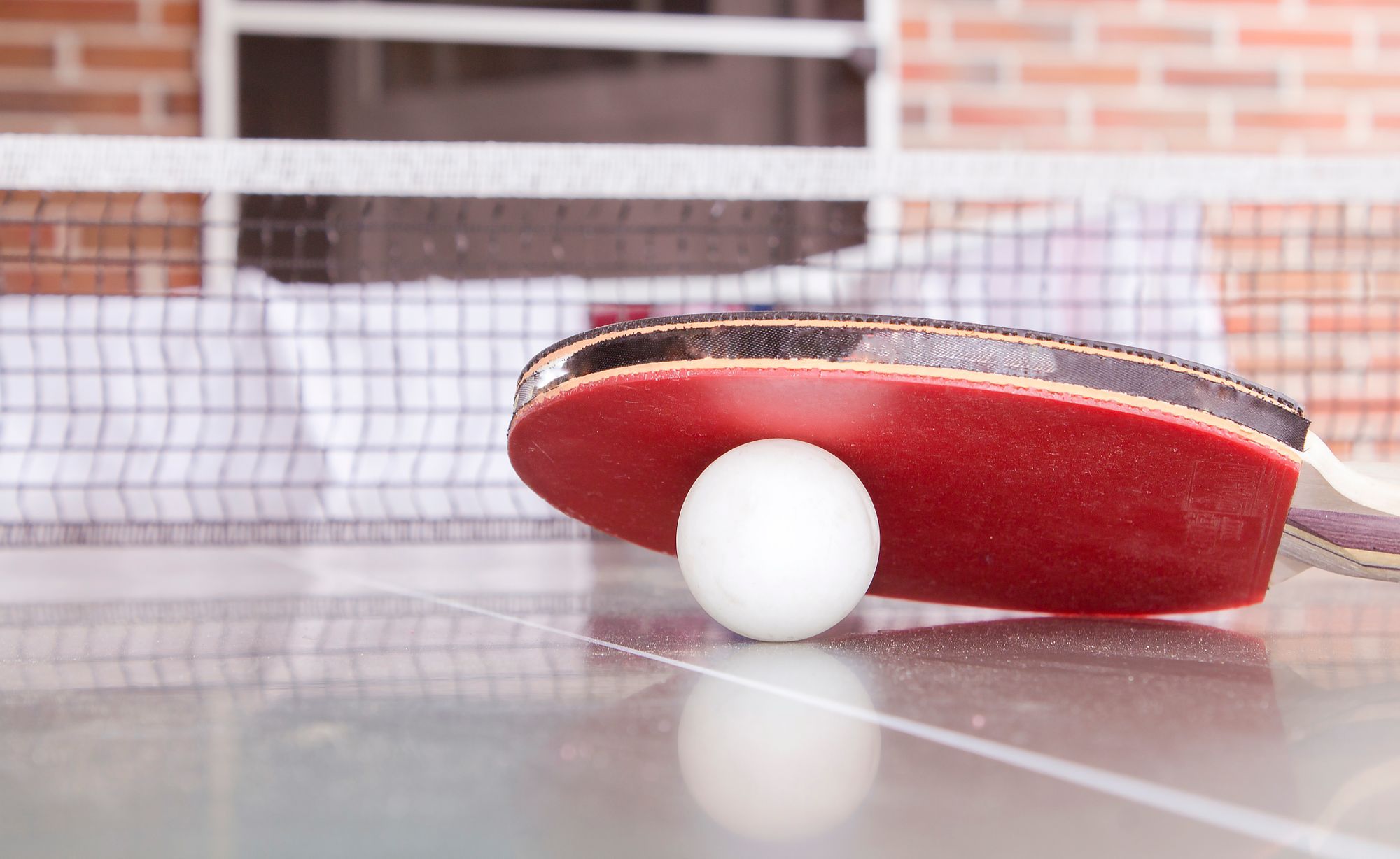 A white table tennis ball resting under the edge of a red bat on a table, with the net behind