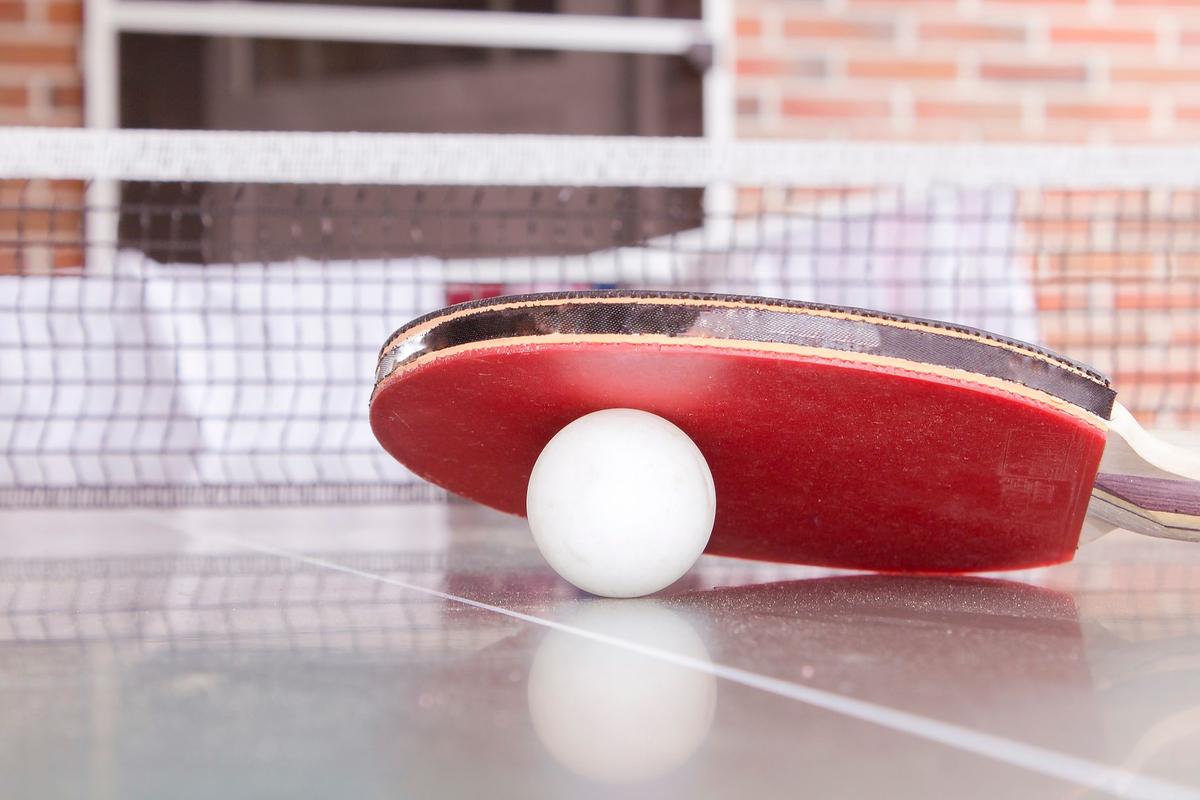 A white table tennis ball resting under the edge of a red bat on a table, with the net behind
