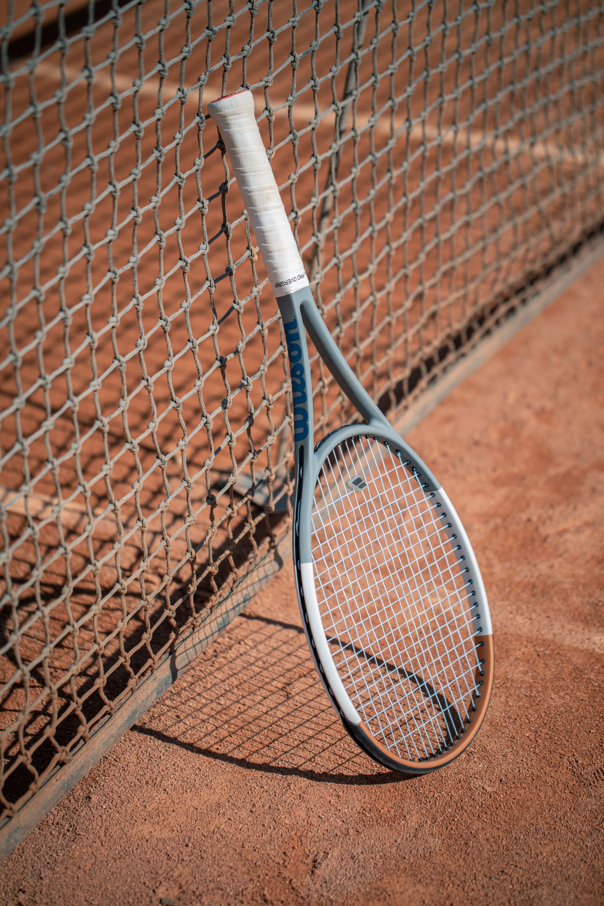 A strung tennis racket leaning against the net on an outdoor clay court