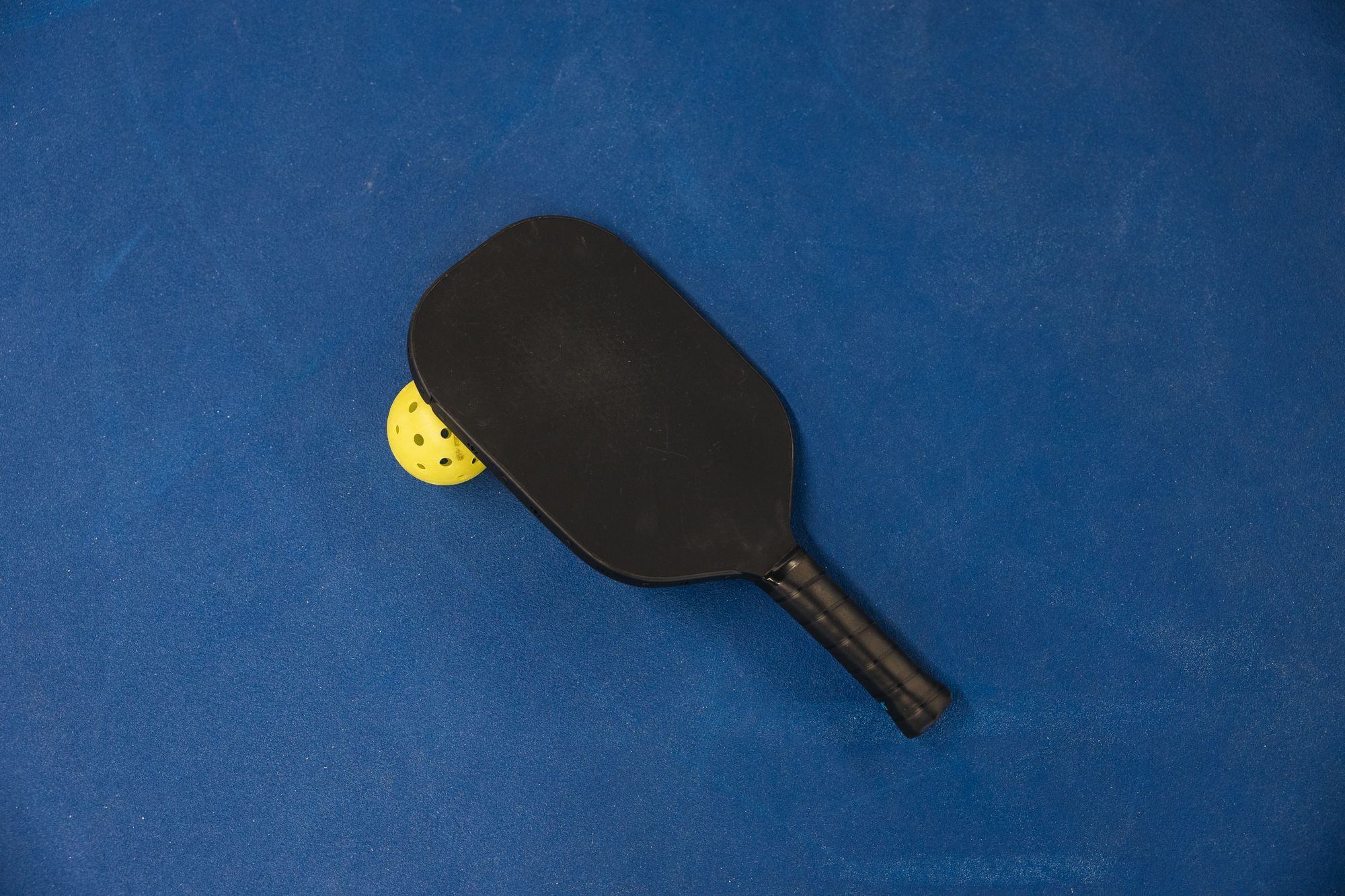 A solid black pickleball paddle lying on a blue court surface, with a yellow holed ball tucked beneath its edge