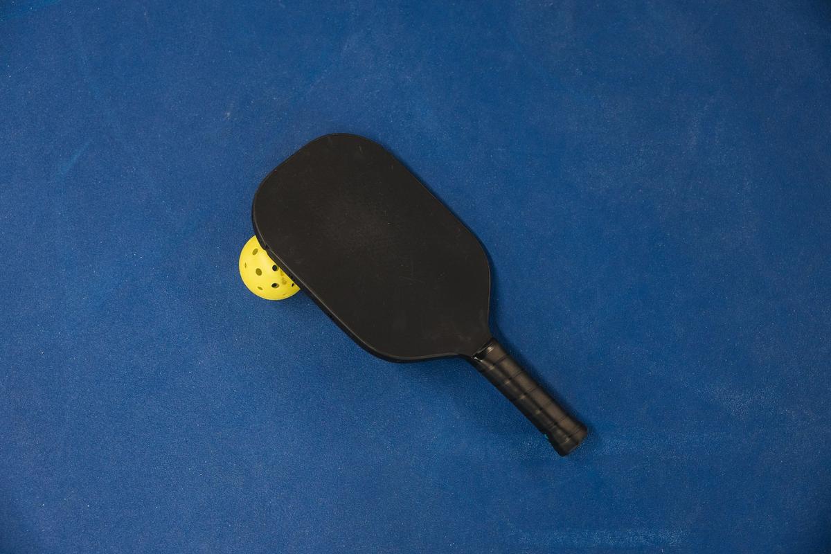 A solid black pickleball paddle lying on a blue court surface, with a yellow holed ball tucked beneath its edge