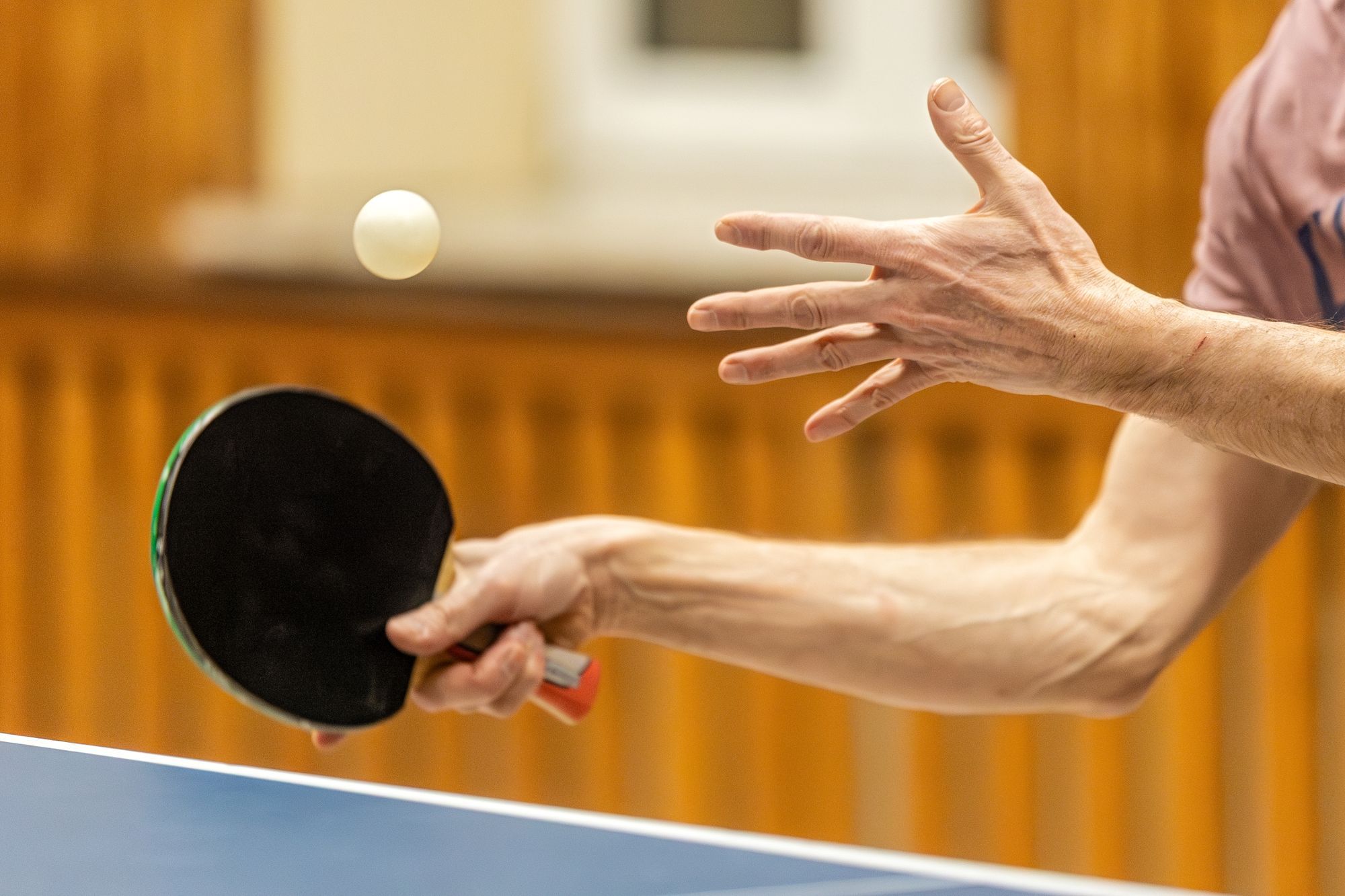 A table tennis player tosses the ball for a serve, bat ready, over the edge of a blue table