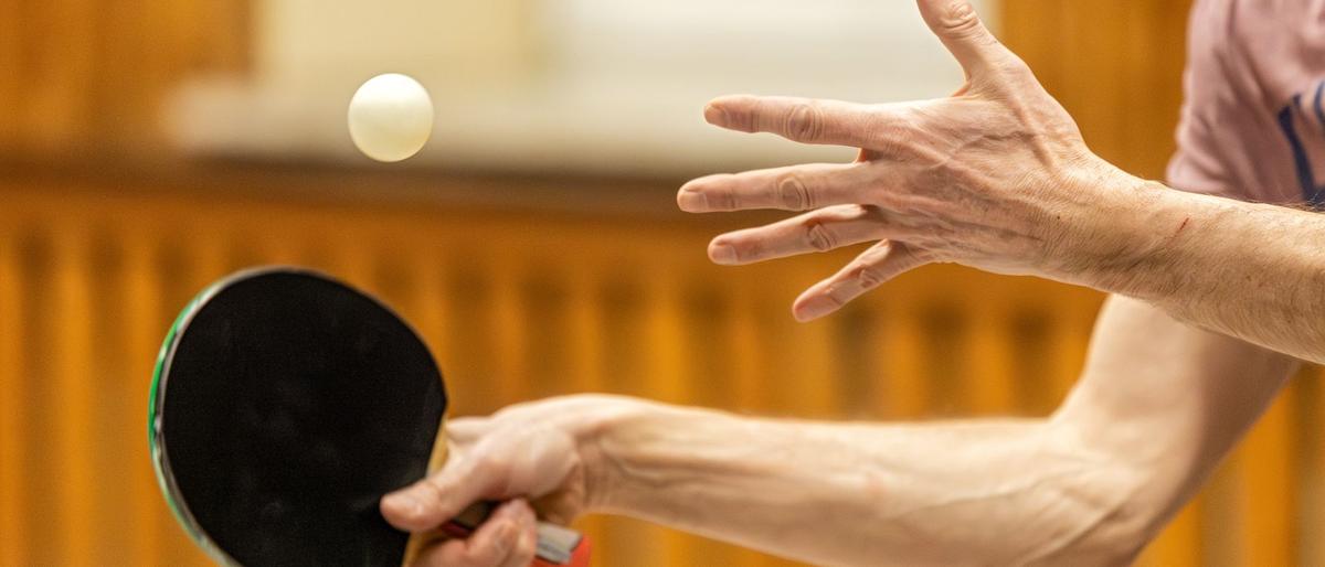 A table tennis player tosses the ball for a serve, bat ready, over the edge of a blue table