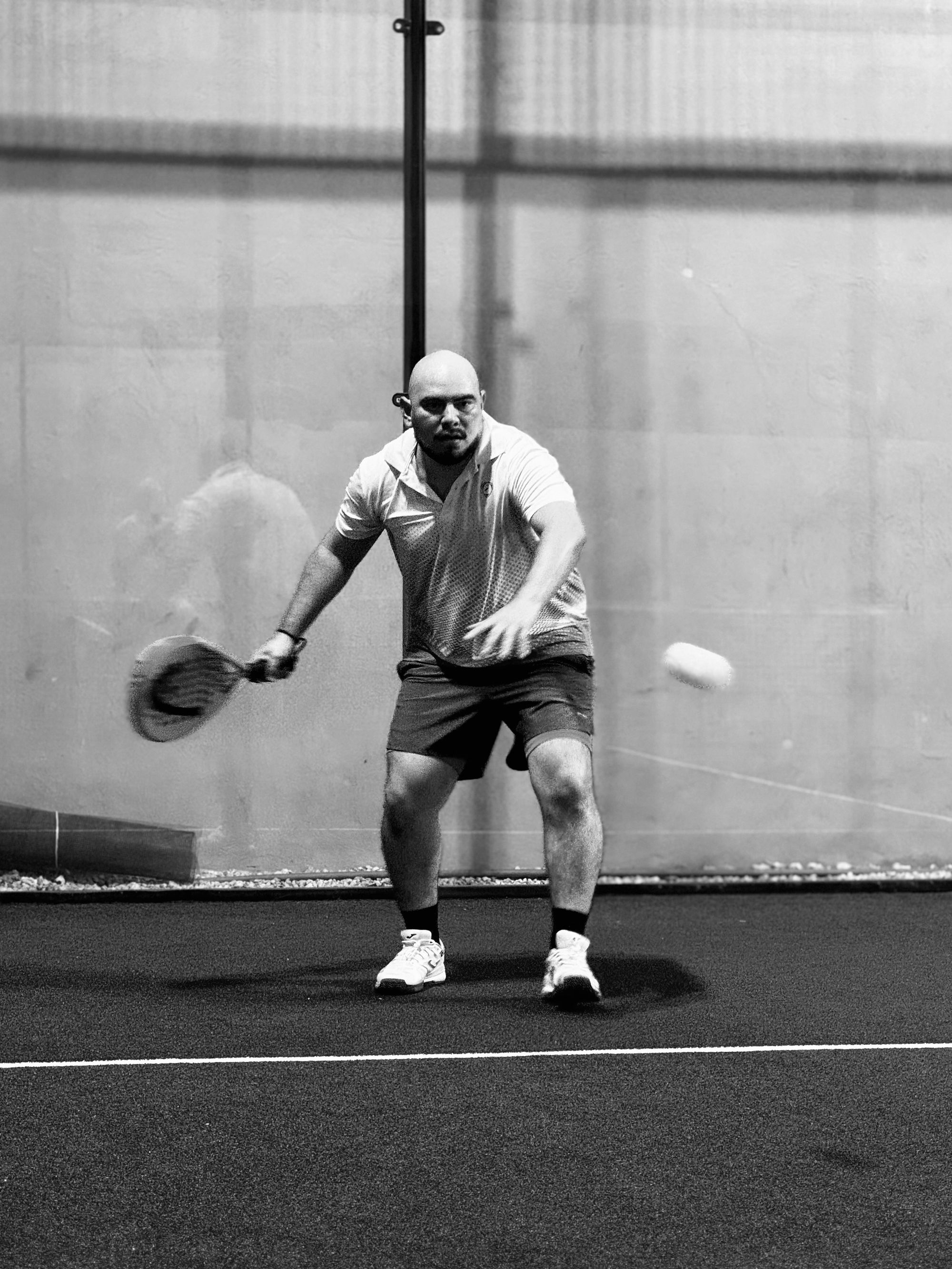 Black-and-white photo of a padel player hitting a low forehand on an indoor court, with the glass back wall behind