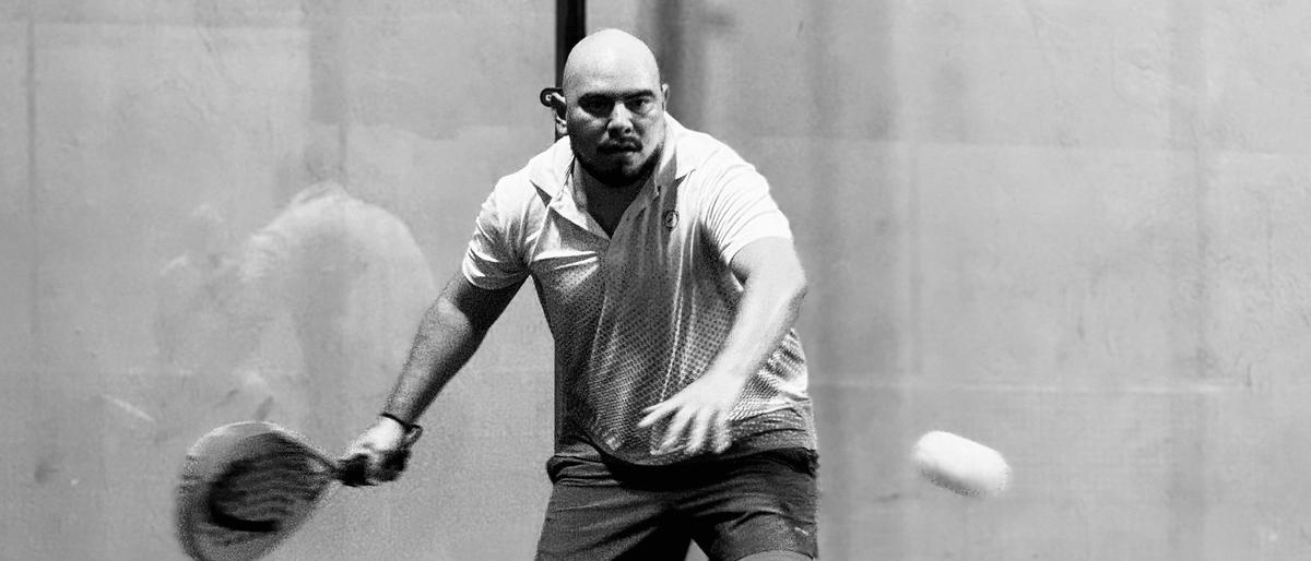 Black-and-white photo of a padel player hitting a low forehand on an indoor court, with the glass back wall behind