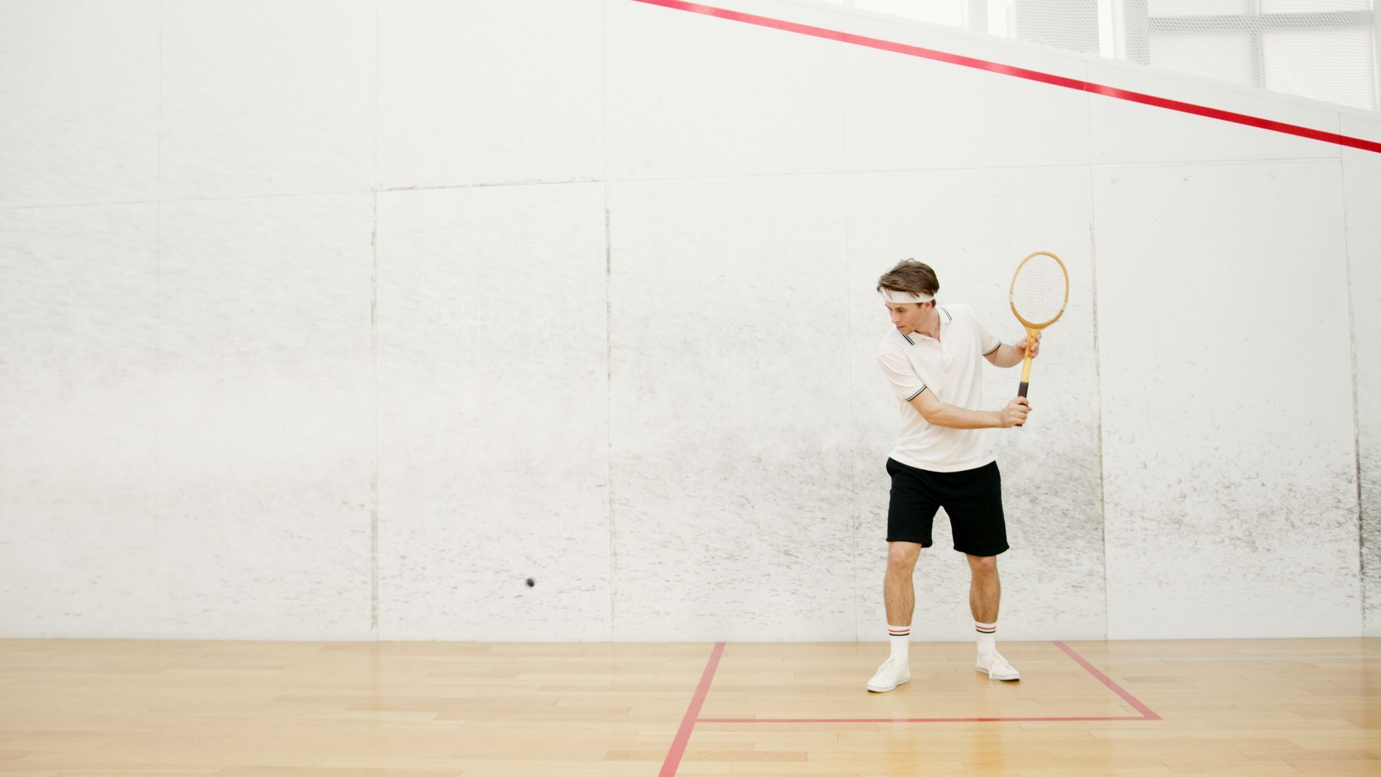 A man in a white shirt and black shorts prepares to swing on an indoor squash court with white walls, red court lines and a wooden floor, with the small black ball in flight to his left