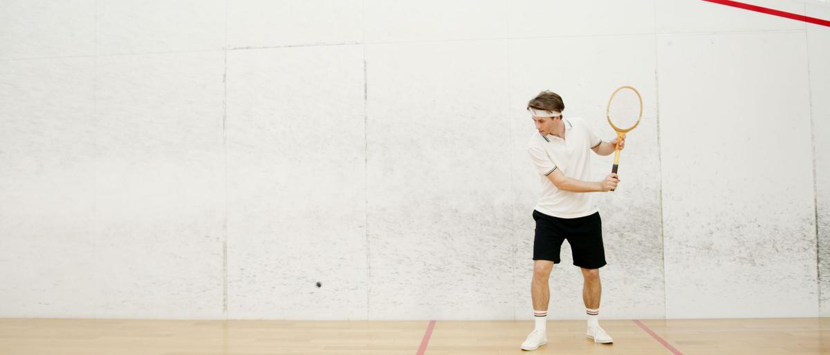 A man in a white shirt and black shorts prepares to swing on an indoor squash court with white walls, red court lines and a wooden floor, with the small black ball in flight to his left
