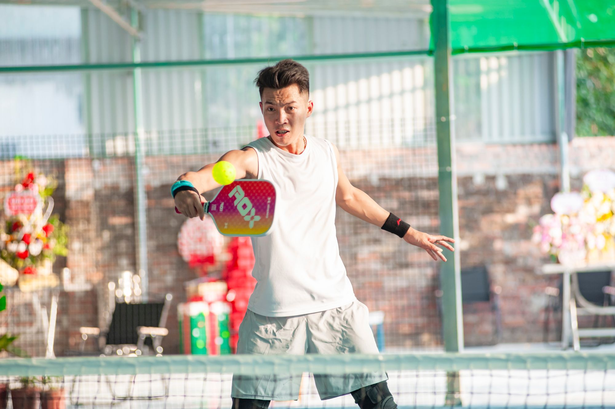 A pickleball player volleys a yellow holed plastic ball with a solid paddle at the net of a covered court