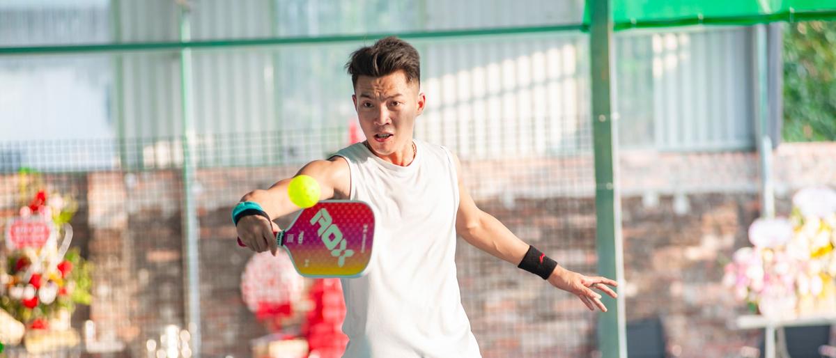 A pickleball player volleys a yellow holed plastic ball with a solid paddle at the net of a covered court